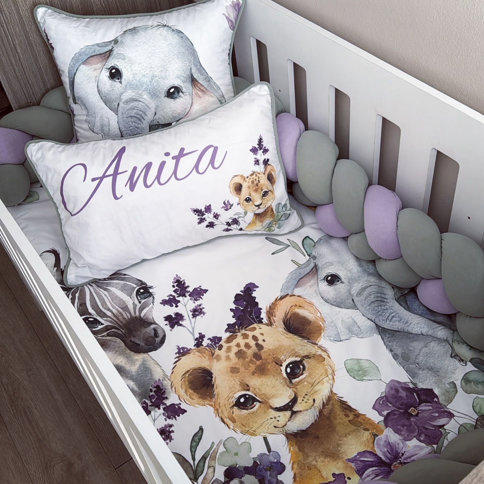 Customizable baby bedding set in a white crib, featuring a watercolor print of an elephant, zebra, lion cub, and purple flowers. Includes a white crib bumper with lavender and gray beads, a personalized pillow reading 'Anita' in purple script, and a matching elephant pillow.
