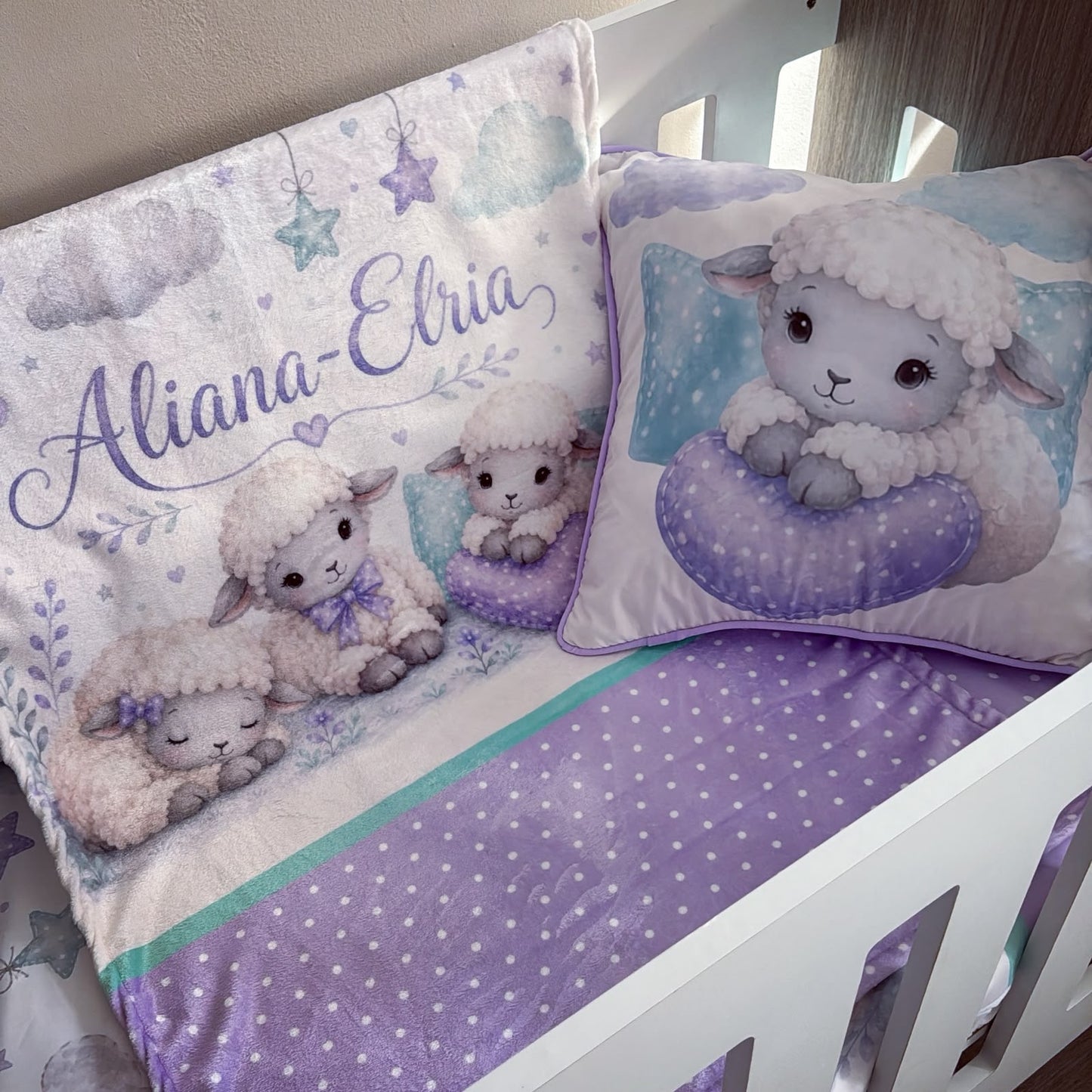 A baby bedding set in a white crib, featuring a blanket and matching pillow with cute cartoon sheep illustrations, purple accents, and the personalized name 'Aliana-Ebria' in purple script.