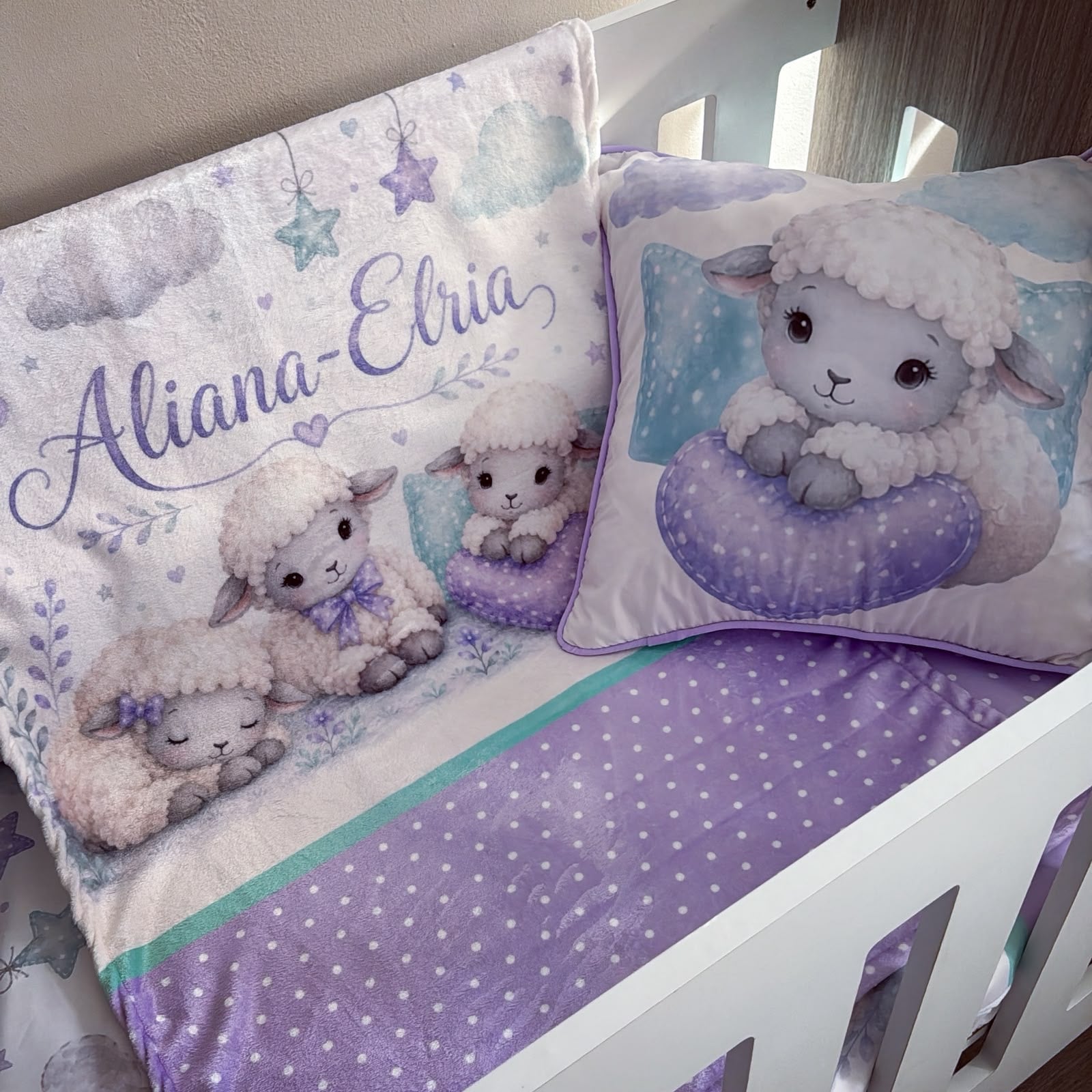 A baby bedding set in a white crib, featuring a blanket and matching pillow with cute cartoon sheep illustrations, purple accents, and the personalized name 'Aliana-Ebria' in purple script.