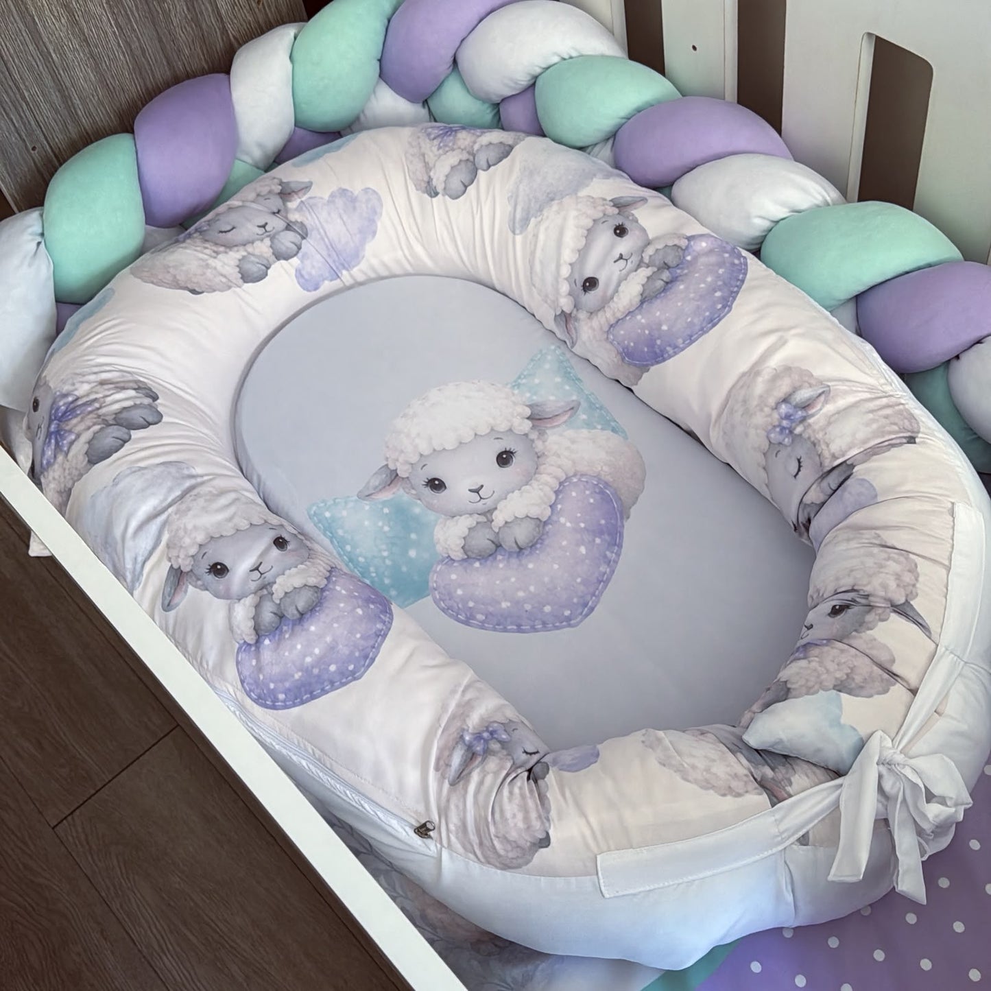 A baby bassinet bedding set featuring a padded bassinet cover with a white background and a lavender and gray lamb pattern, a matching oval insert with a central lamb illustration, and a braided crib bumper in pastel purple, mint green, and white.