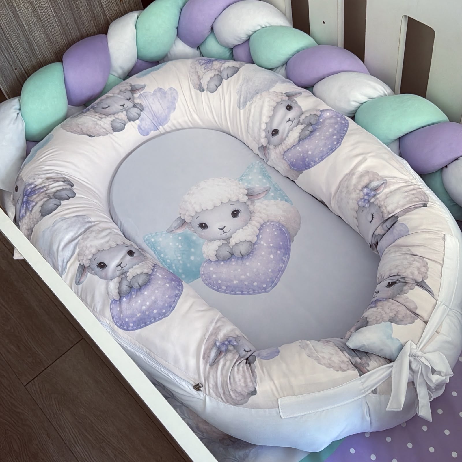 A baby bassinet bedding set featuring a padded bassinet cover with a white background and a lavender and gray lamb pattern, a matching oval insert with a central lamb illustration, and a braided crib bumper in pastel purple, mint green, and white.
