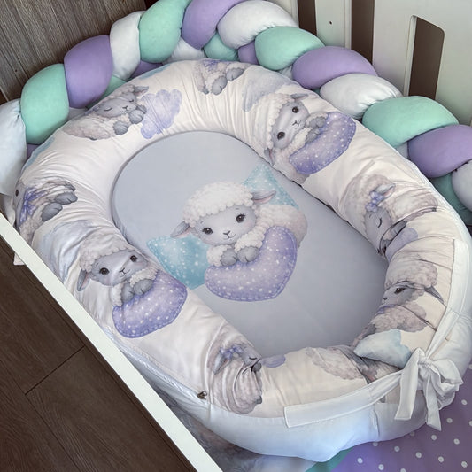 A baby bassinet bedding set featuring a padded bassinet cover with a white background and a lavender and gray lamb pattern, a matching oval insert with a central lamb illustration, and a braided crib bumper in pastel purple, mint green, and white.
