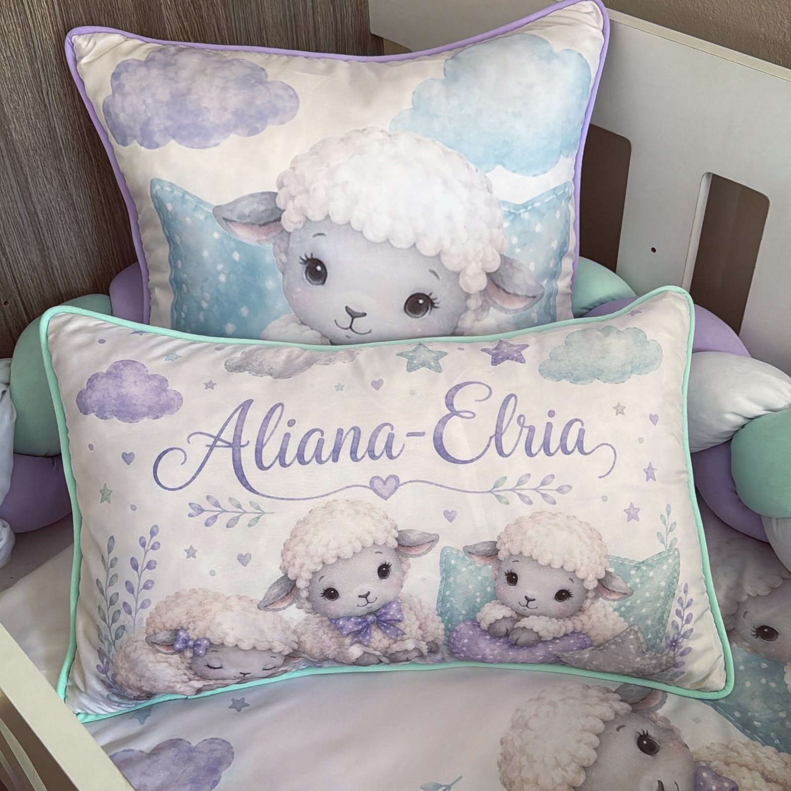Personalized baby bedding set featuring two plush throw pillows in a nursery crib. The front pillow is personalized with the name 'Aliana-Elria' in purple script, and the back pillow shows a cute cartoon lamb.