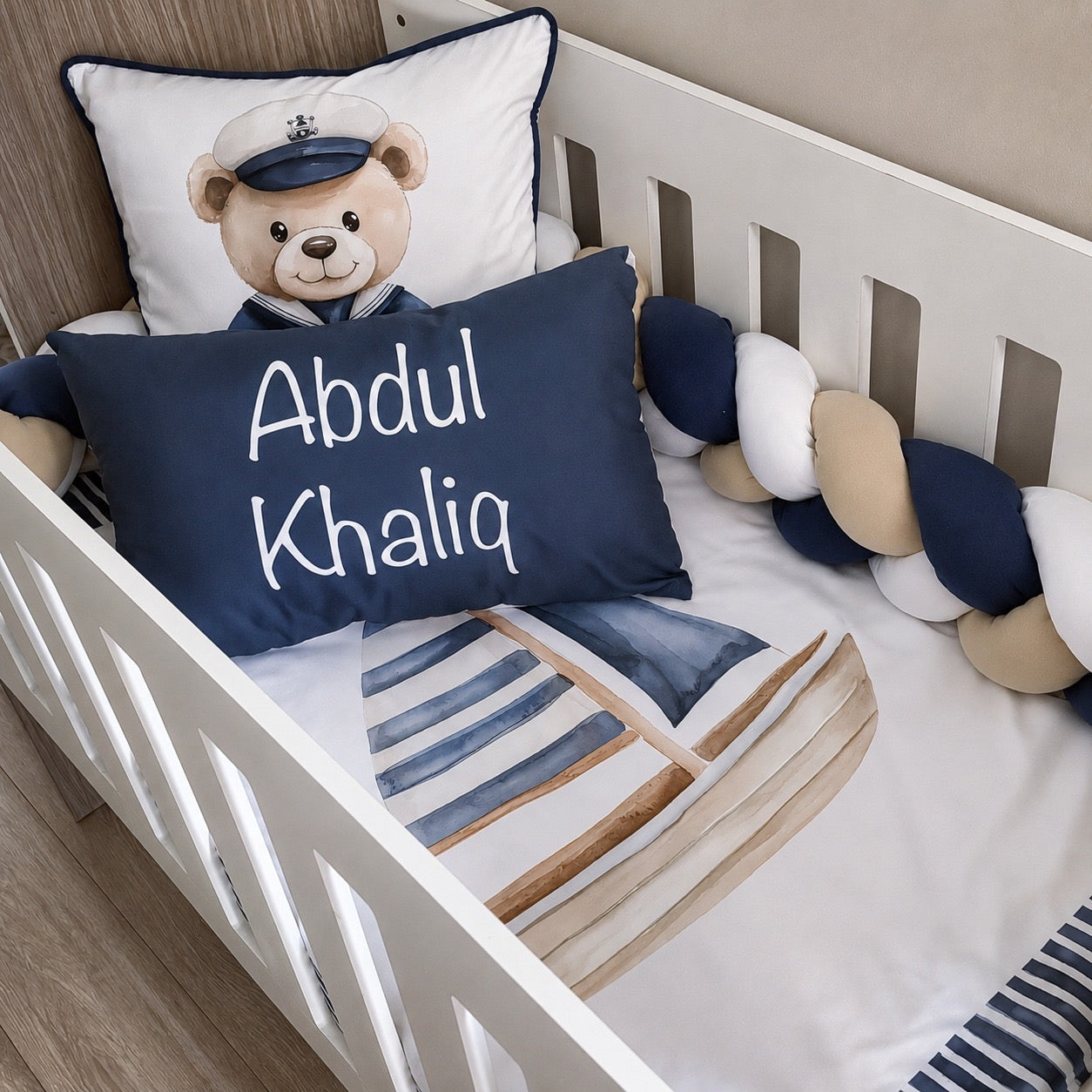 A personalized baby crib bedding set featuring a white crib with a watercolor sailboat print, a navy blue personalized pillow reading 'Abdul Khaliq', a white pillow with a teddy bear wearing a captain hat, and a chunky braided bumper in navy, white, and beige.