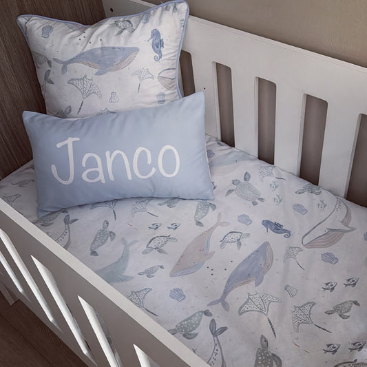A baby bedding set in a white crib, featuring a white duvet cover and pillowcase with a blue-gray sea life print, and a light blue rectangular pillow with the name 'Janco' printed in white.