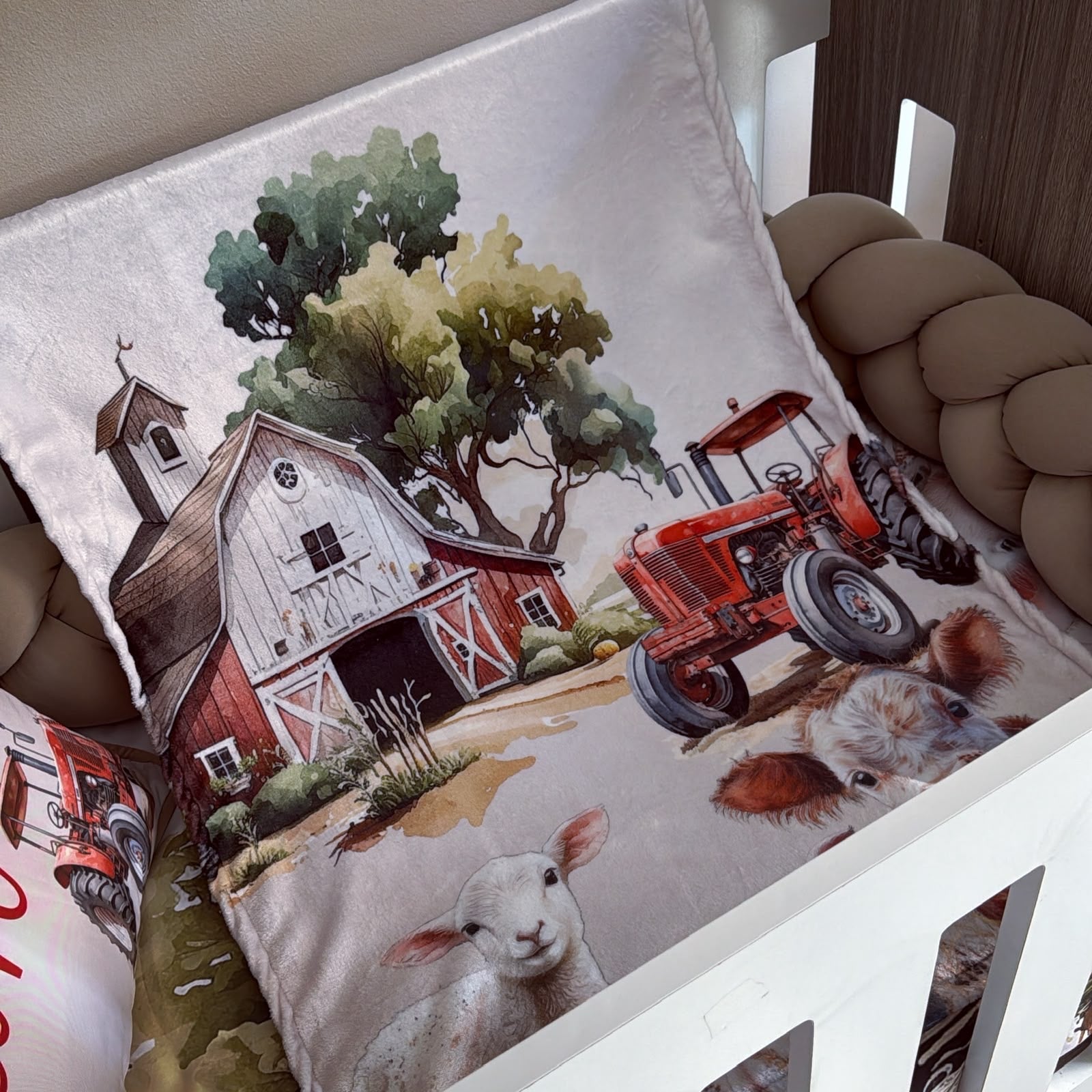 A printed baby blanket or quilt featuring a farm scene with a red tractor, barn, trees, and animals, styled inside a white crib with a chunky beige cord bumper.