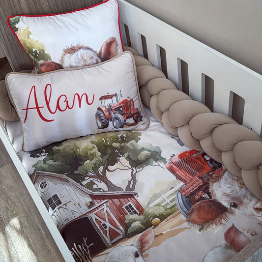 A personalized baby crib bedding set featuring a farm-themed print with a red tractor, barn, trees, and a cow, complete with a matching braided bumper and a custom name pillow reading 'Alan'.