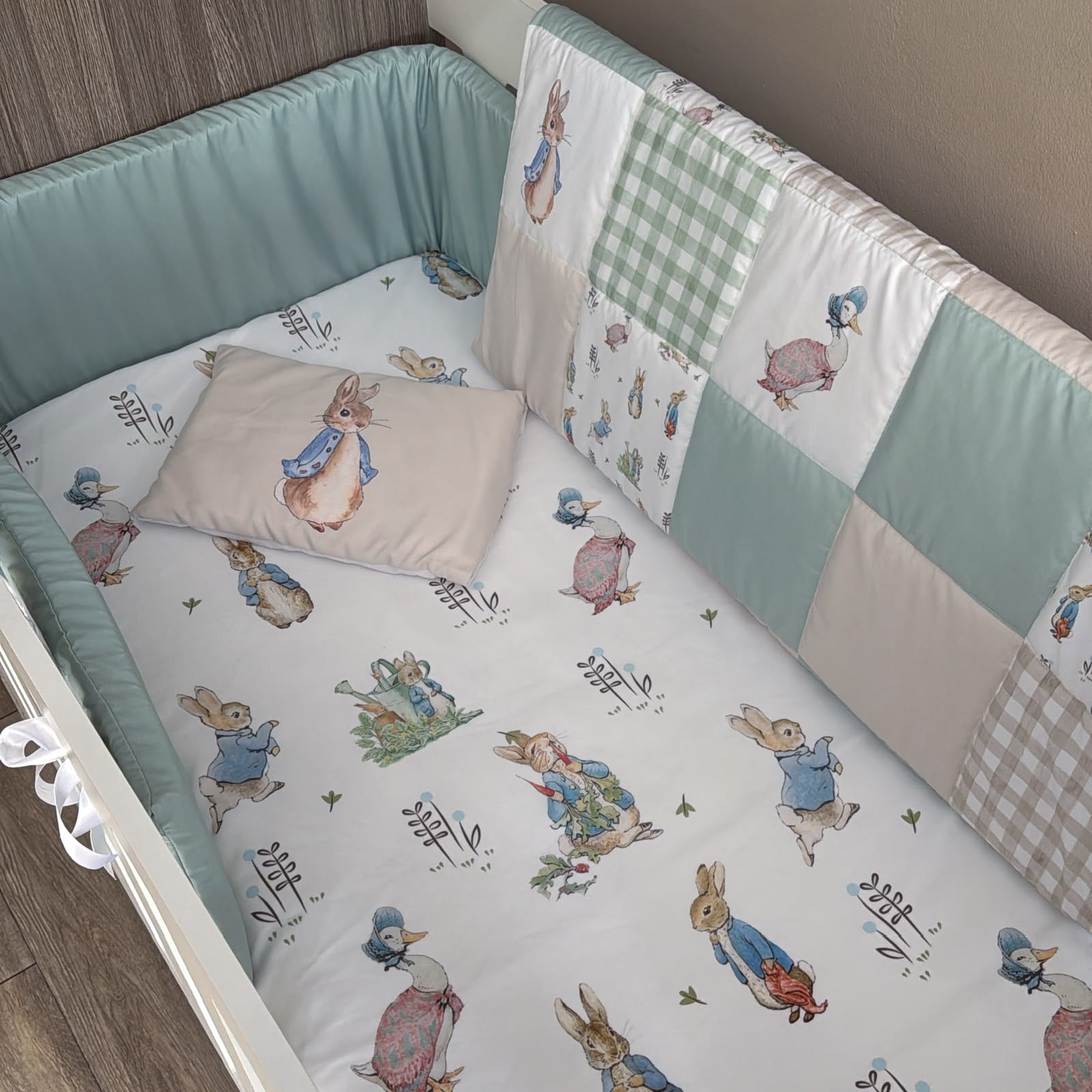 A baby crib bedding set featuring a patchwork quilted bumper with Peter Rabbit illustrations, a matching crib sheet, and a soft cushion pillow, shown in a crib.