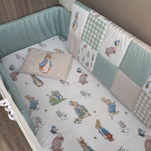 A baby crib bedding set featuring a patchwork quilted bumper with Peter Rabbit illustrations, a matching crib sheet, and a soft cushion pillow, shown in a crib.