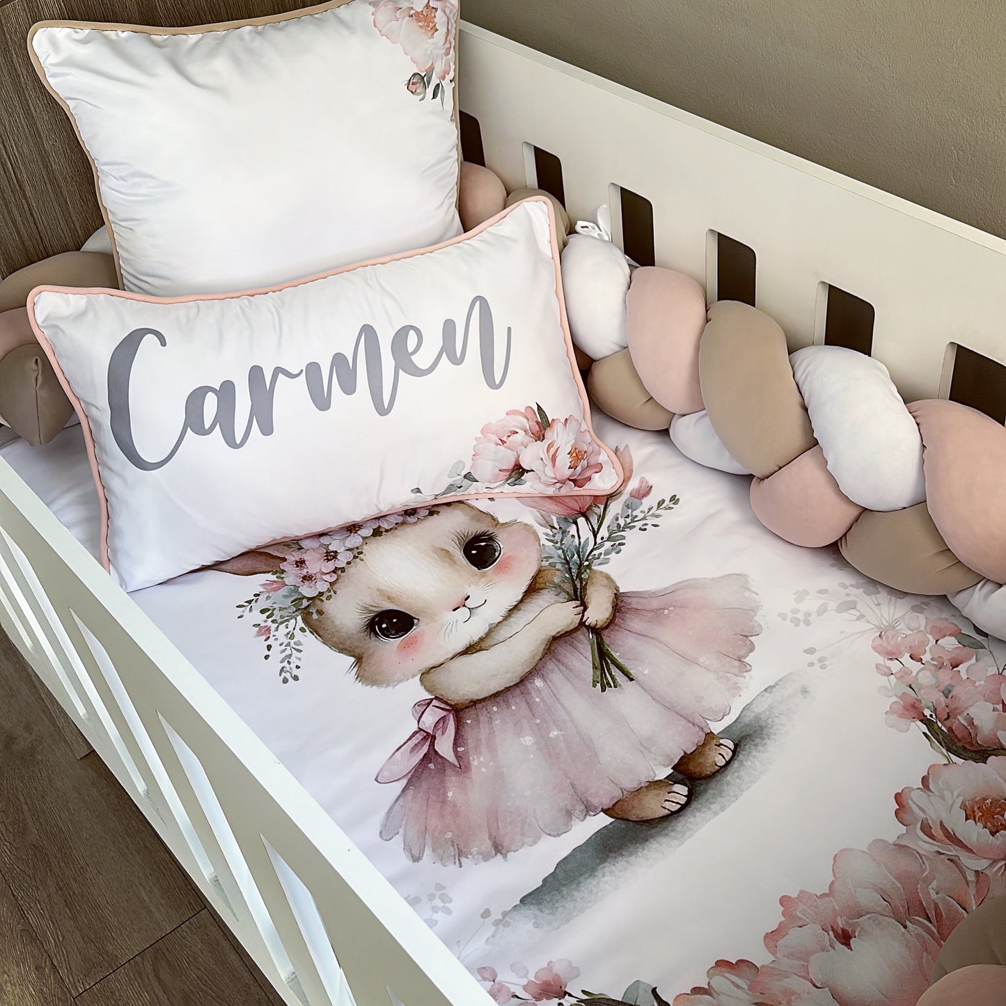 A cot bedding set featuring a pink bunny design, with a personalized name pillow, a duvet cover, and a fitted sheet, arranged on a white cot.