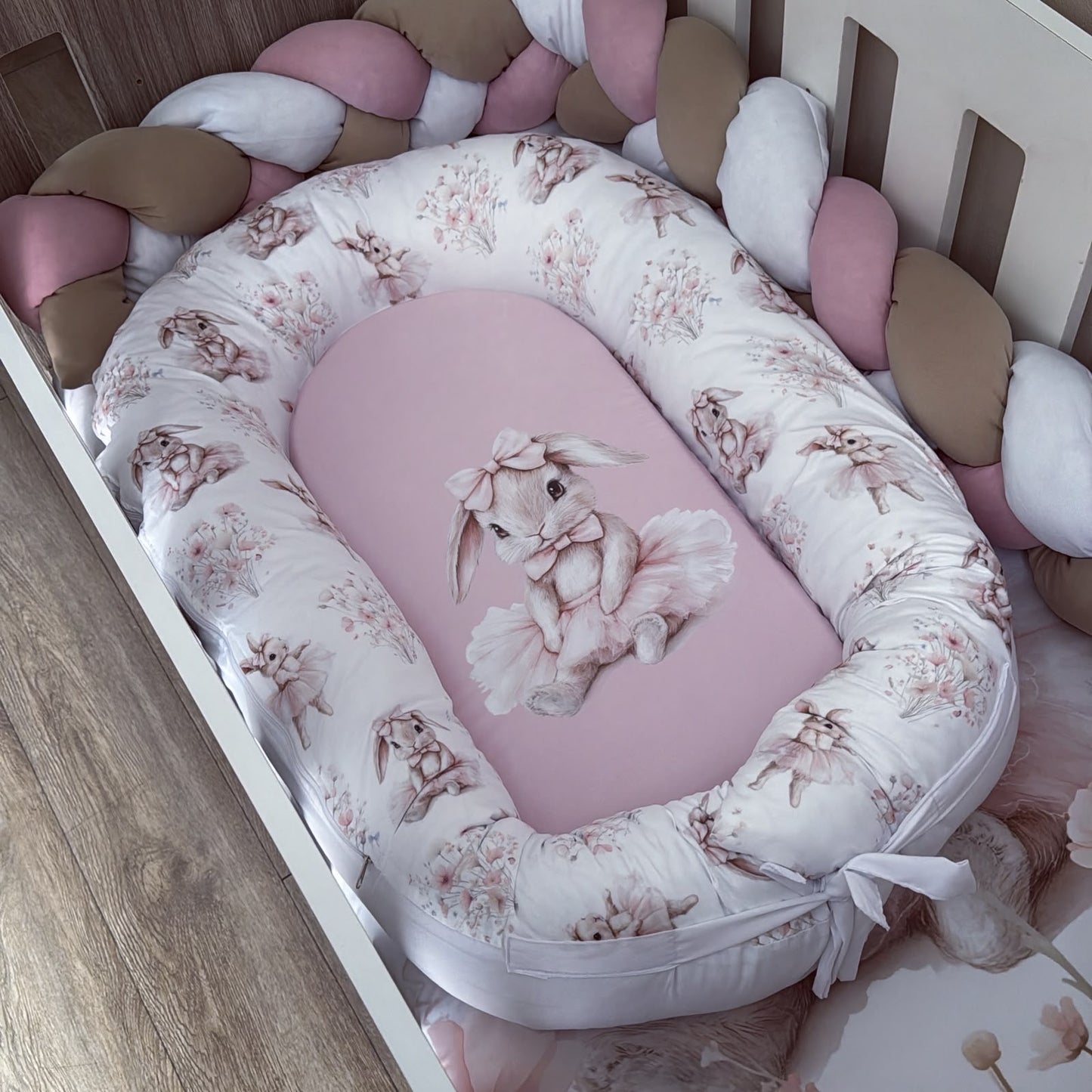 A baby bassinet bedding set featuring a pink bunny-themed mattress cover and a white bumper pad with a dusty pink bunny and floral print, styled inside a white wooden crib with braided pink and beige crib pillows.