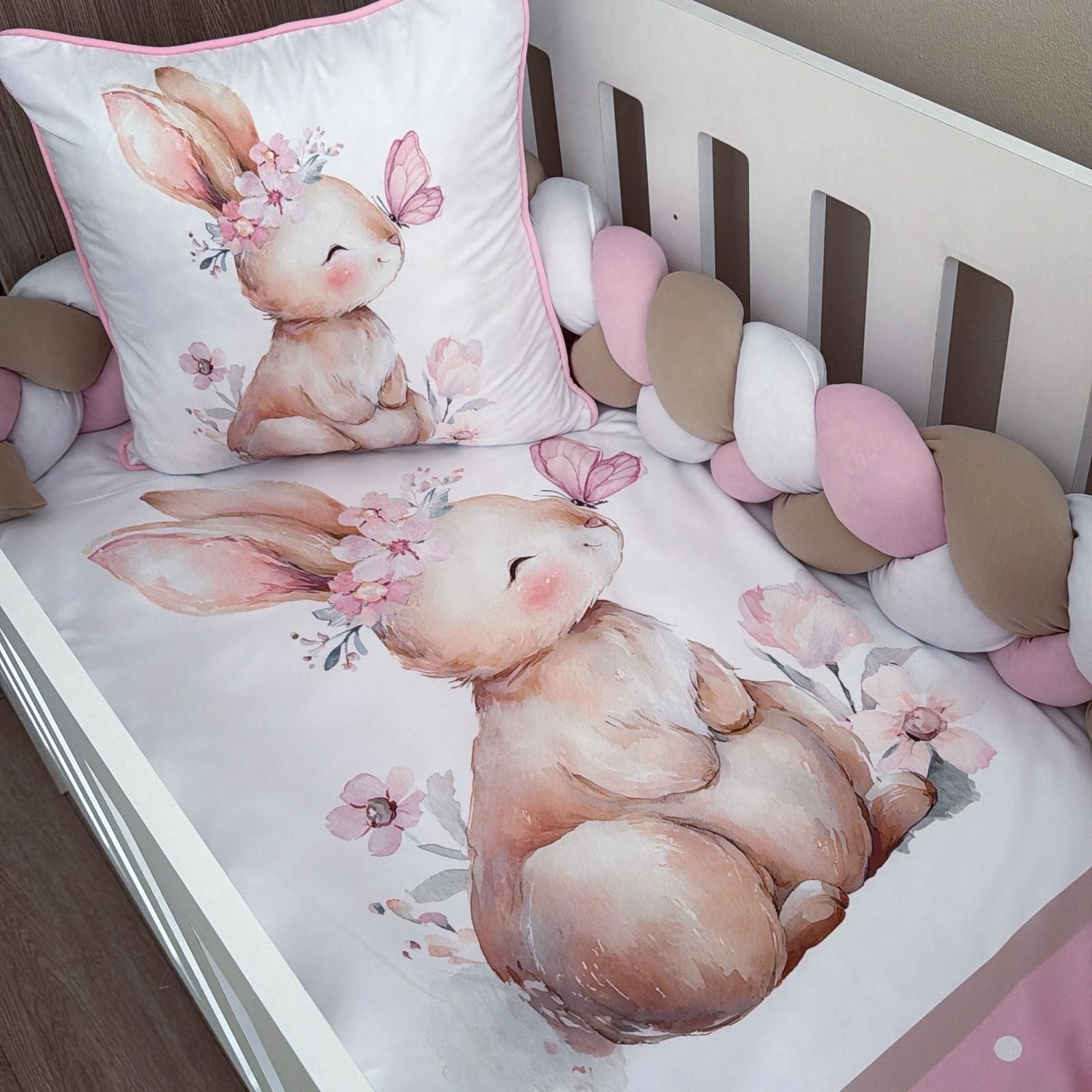 A white crib bedding set featuring a watercolor-style bunny illustration with pink flowers and a butterfly, including a fitted sheet, pillowcase, and chunky crib bumper in soft pink, beige, and white.