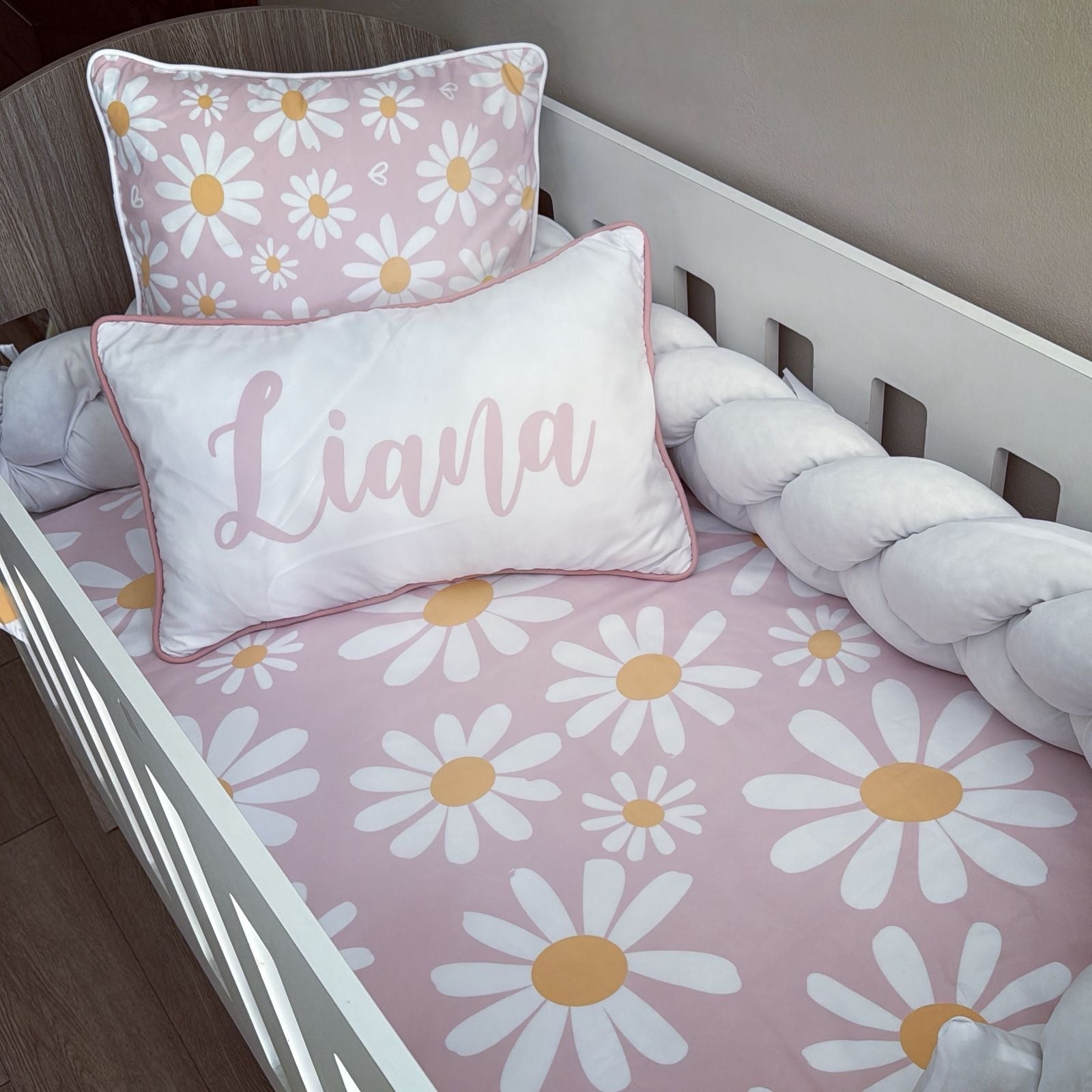 Children's crib with pink daisy pattern bedding and a pillow with 'Liana' on it.