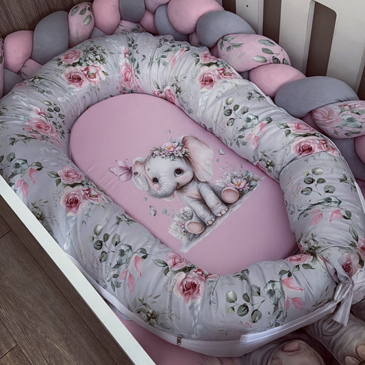 A baby bassinet bedding set featuring a padded bassinet bumper with a pink and gray floral print, a matching pink insert, and a central elephant cushion, shown inside a crib.