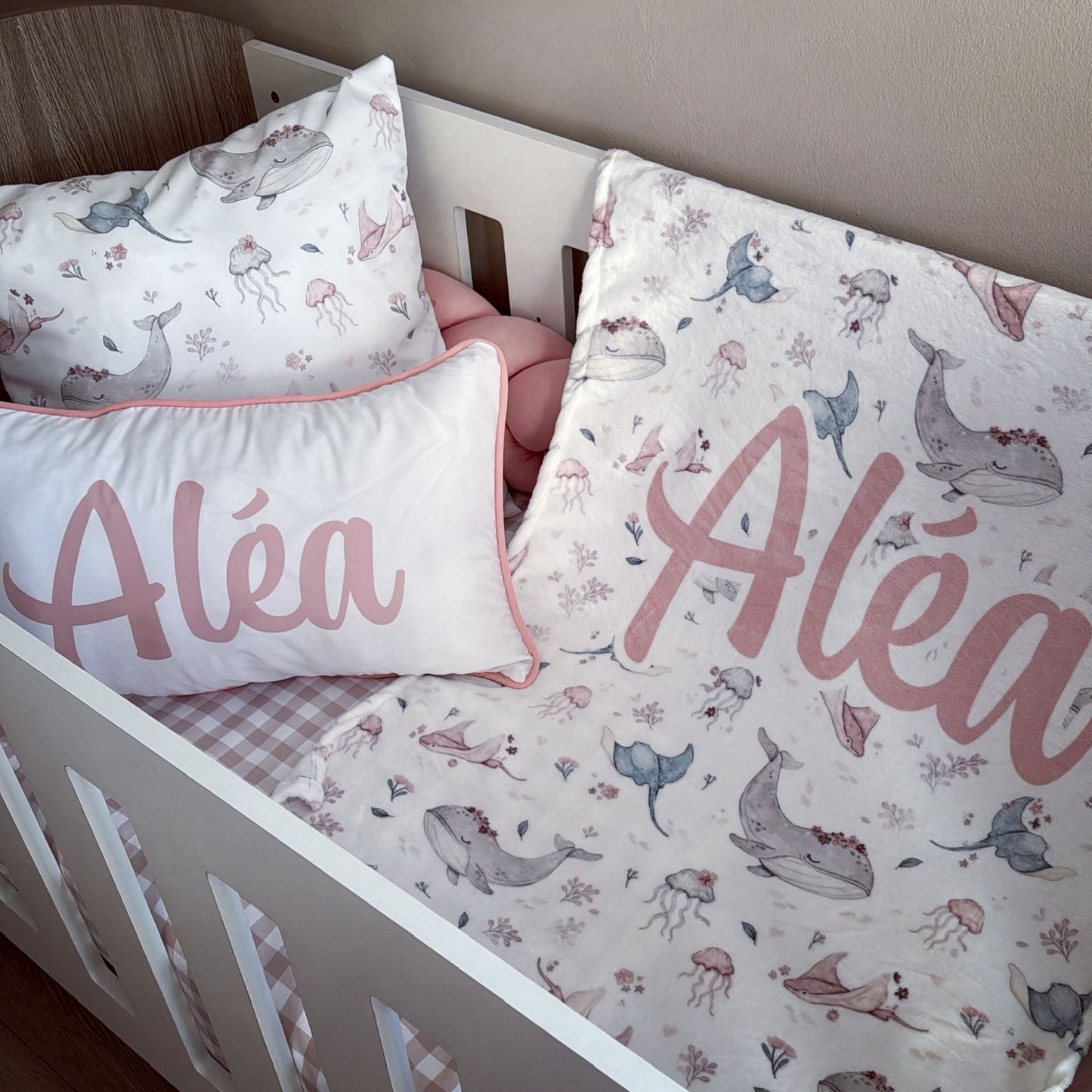 A personalized baby bedding set in a white wooden crib, featuring a whale and jellyfish print with the name 'Aléa' printed in pink on two pillows.