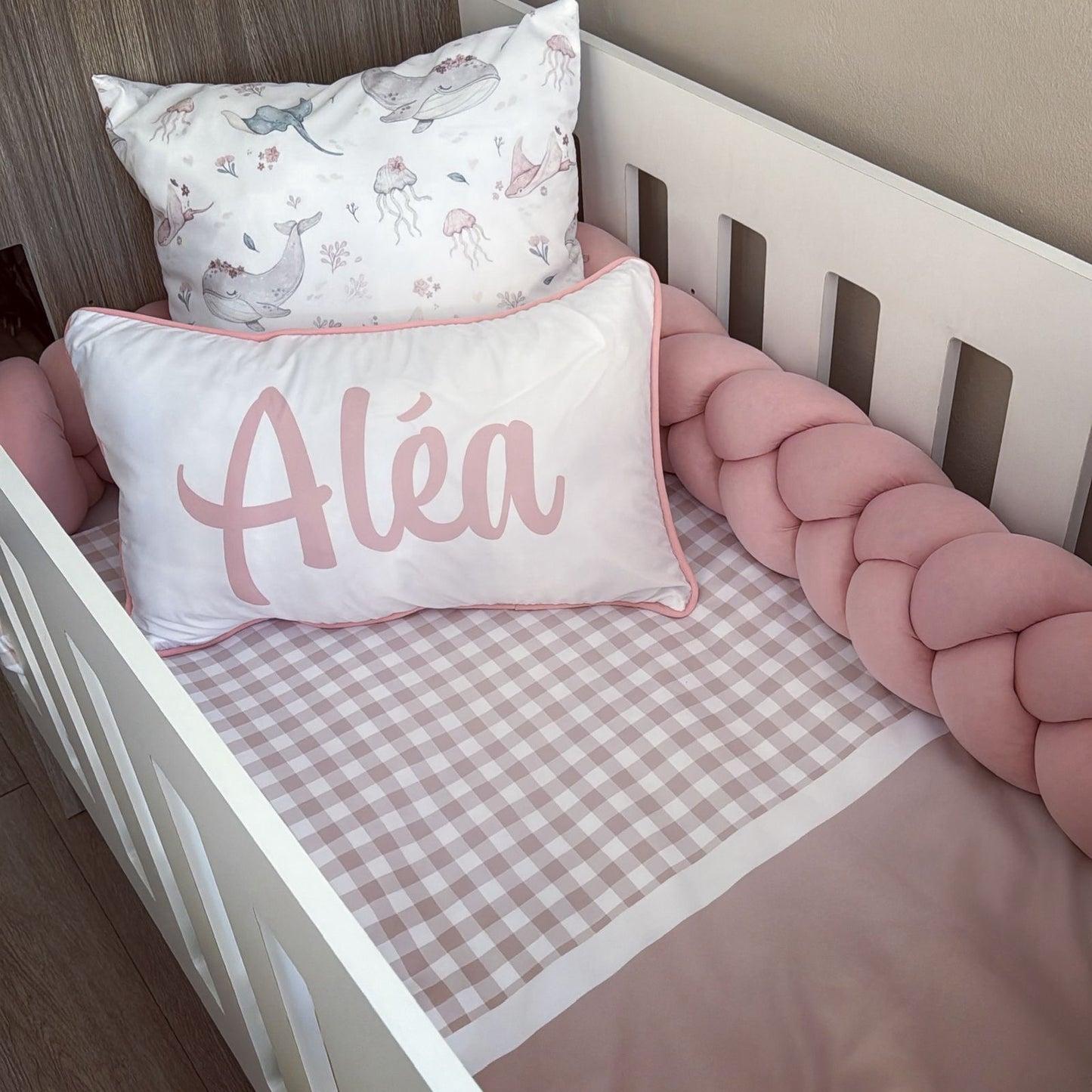 A personalized baby bedding set in a white crib, featuring a pink and white gingham sheet, a pink braided bumper, a white pillow with pink trim and the name 'Aléa' in pink script, and a white pillow with a pastel animal print.