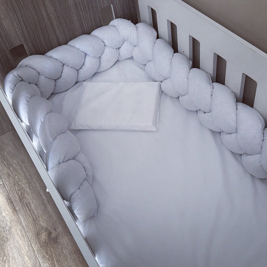White braided crib bumper pad arranged around a white baby crib with white bedding and a matching folded pillow, shown in a nursery setting with wood flooring and wall visible.