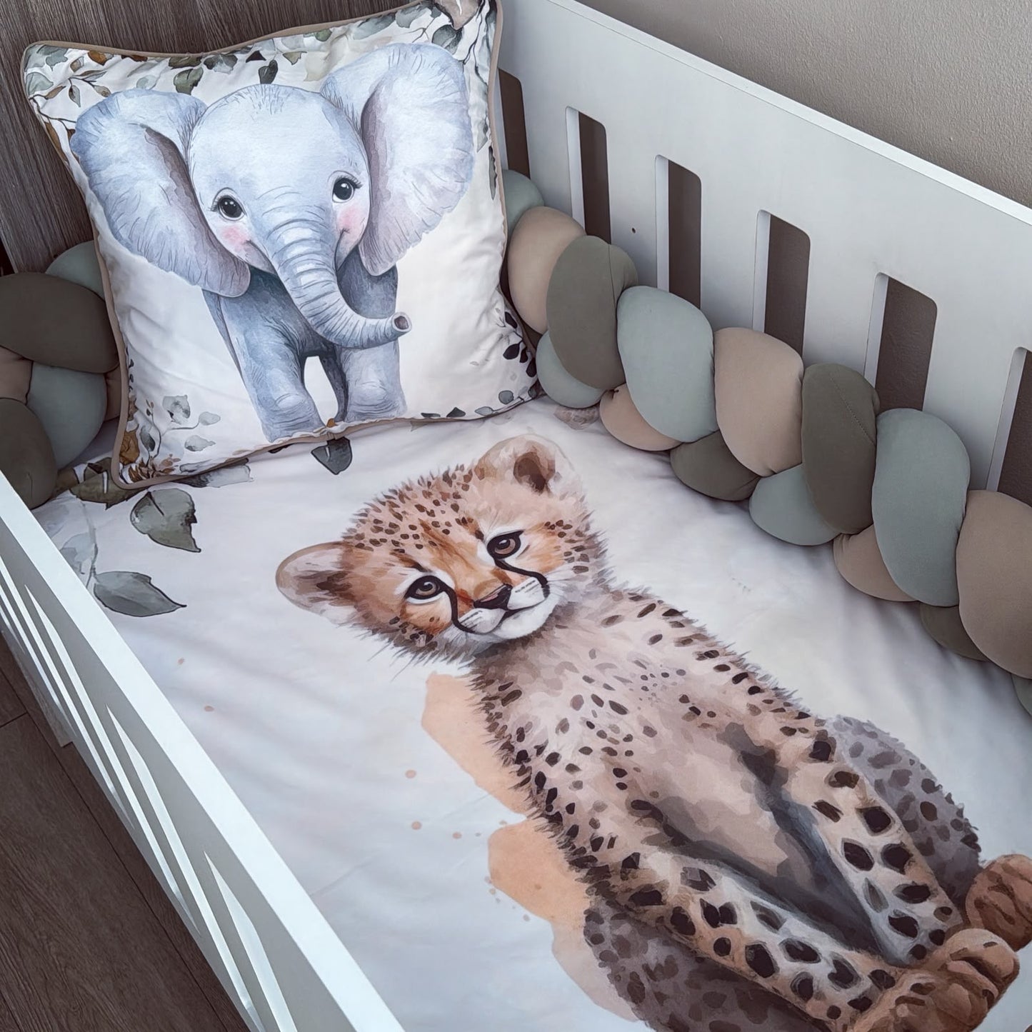A white crib with a watercolor print bedding set featuring a cheetah cub on the mattress and a matching elephant pillow, surrounded by a multi-colored chunky bumper.