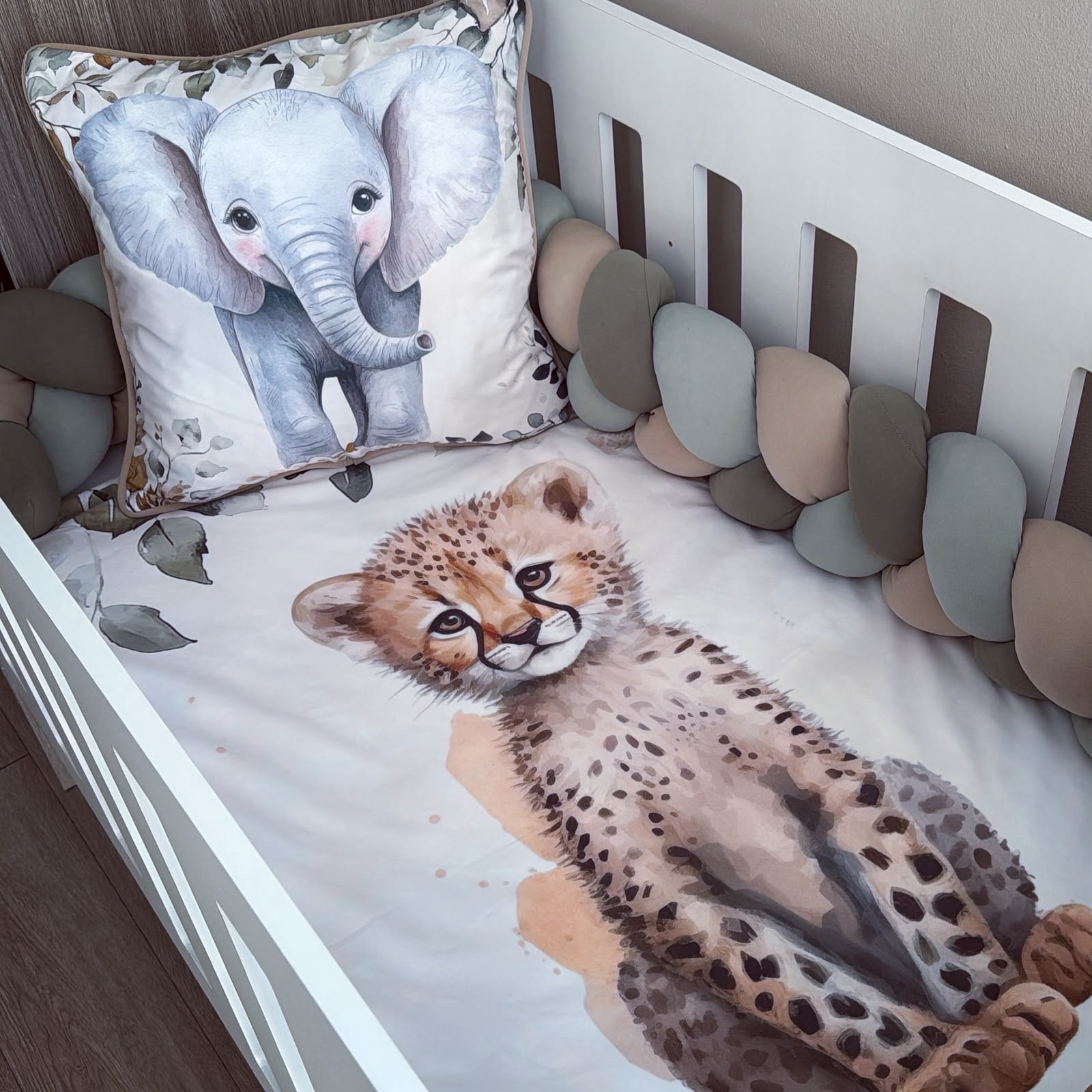 A white crib with a watercolor print bedding set featuring a cheetah cub on the mattress and a matching elephant pillow, surrounded by a multi-colored chunky bumper.