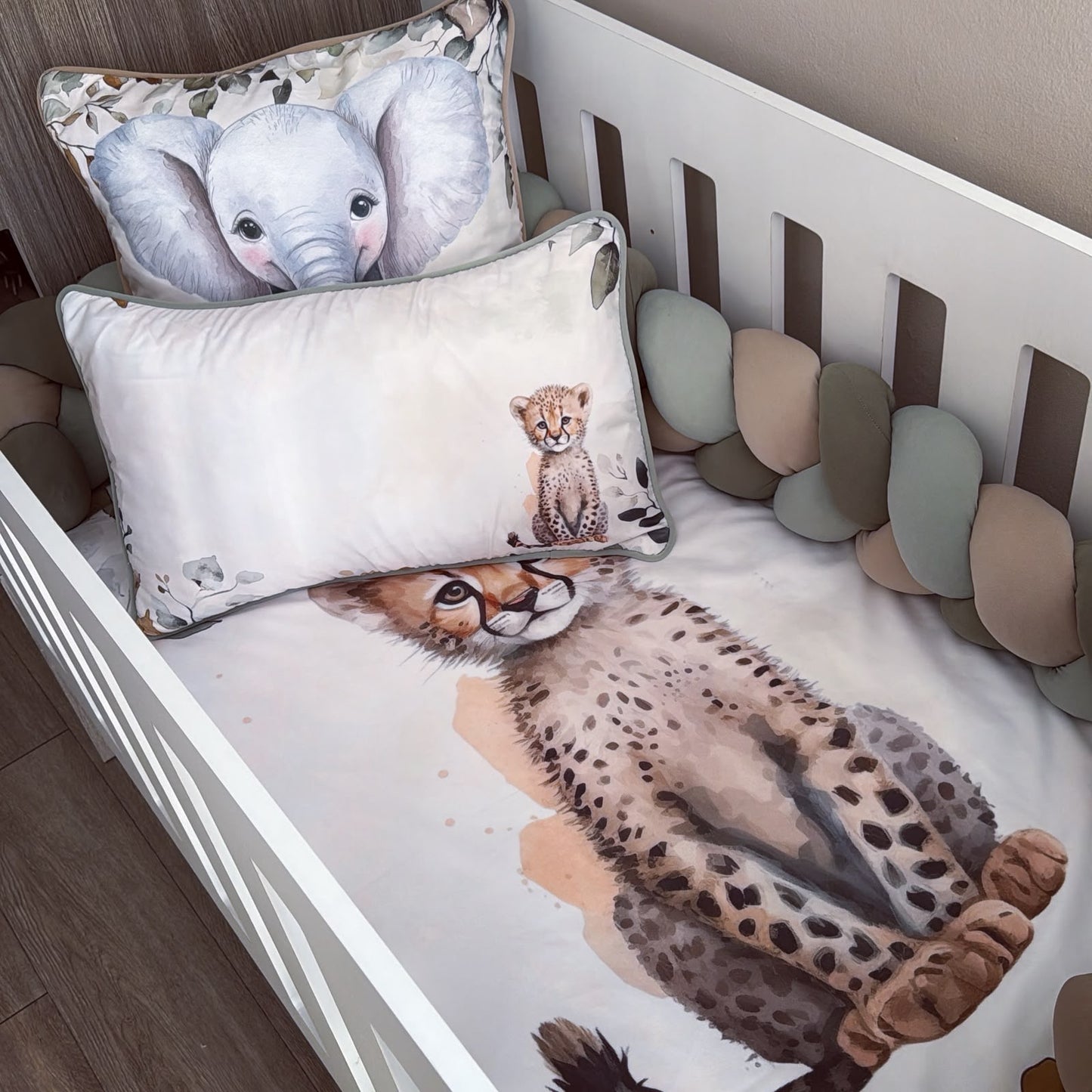 A baby crib bedding set featuring a printed cheetah on the fitted sheet and matching elephant pillowcase, styled inside a white wooden crib with a soft cushion bumper.