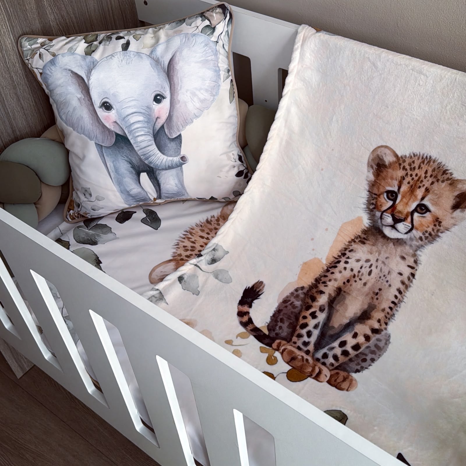 A baby bedding set in a white crib, featuring a cream-colored blanket with a watercolor cheetah print and a matching elephant pillow.