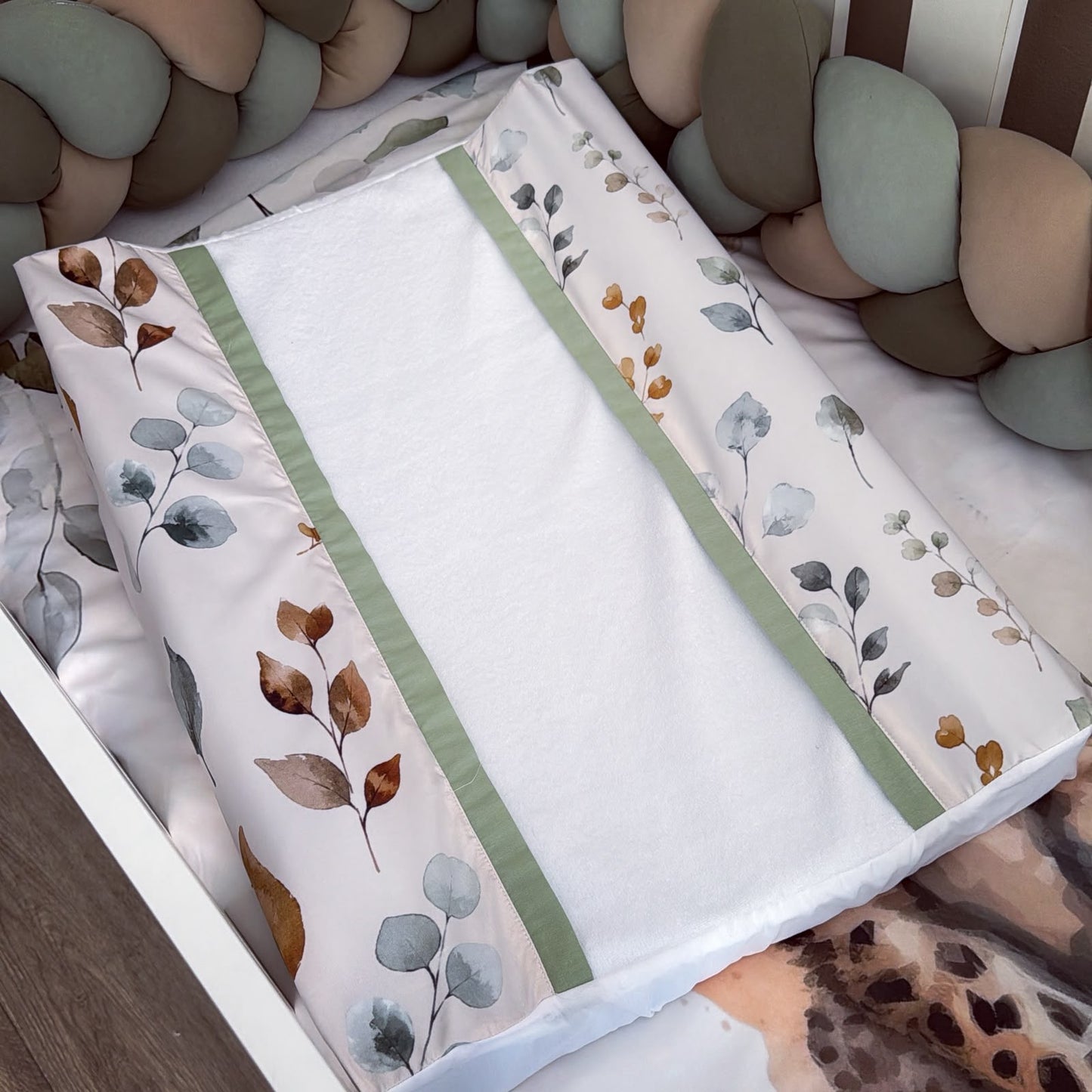 A baby changing pad with a white center and a printed border featuring eucalyptus leaves in muted green, brown, and gray tones, styled in a nursery crib setting.