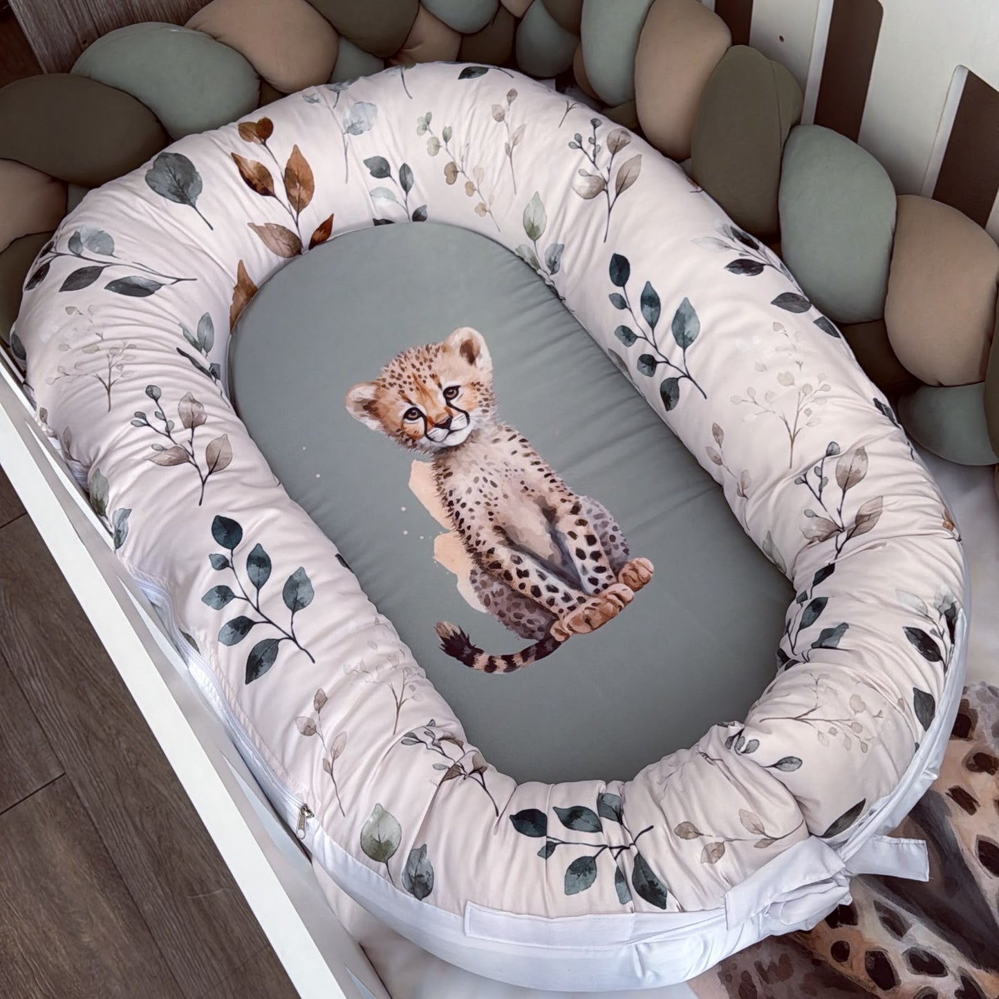 A baby bassinet with a padded oval mattress and a padded bumper pad. The mattress is gray with a printed cheetah cub design, and the bumper pad is cream with a leafy pattern in muted green, brown, and gray.