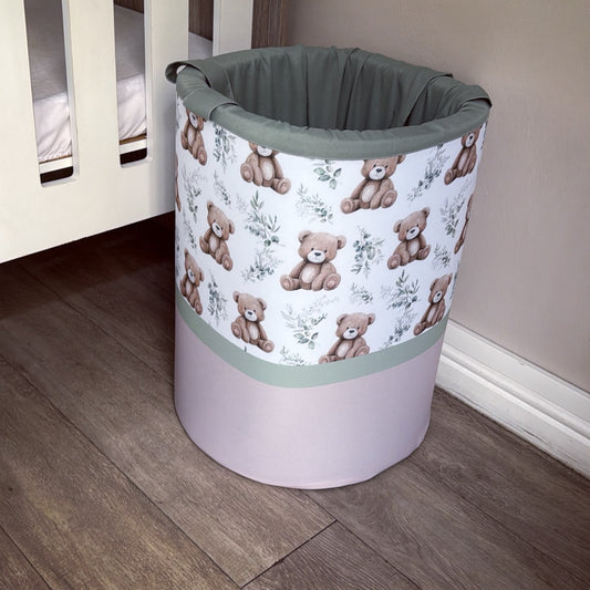 A fabric cylindrical laundry hamper with a white upper section printed with brown teddy bears and green floral motifs, a sage green top rim, and a solid pale pink lower half, shown on a wood floor next to a crib.