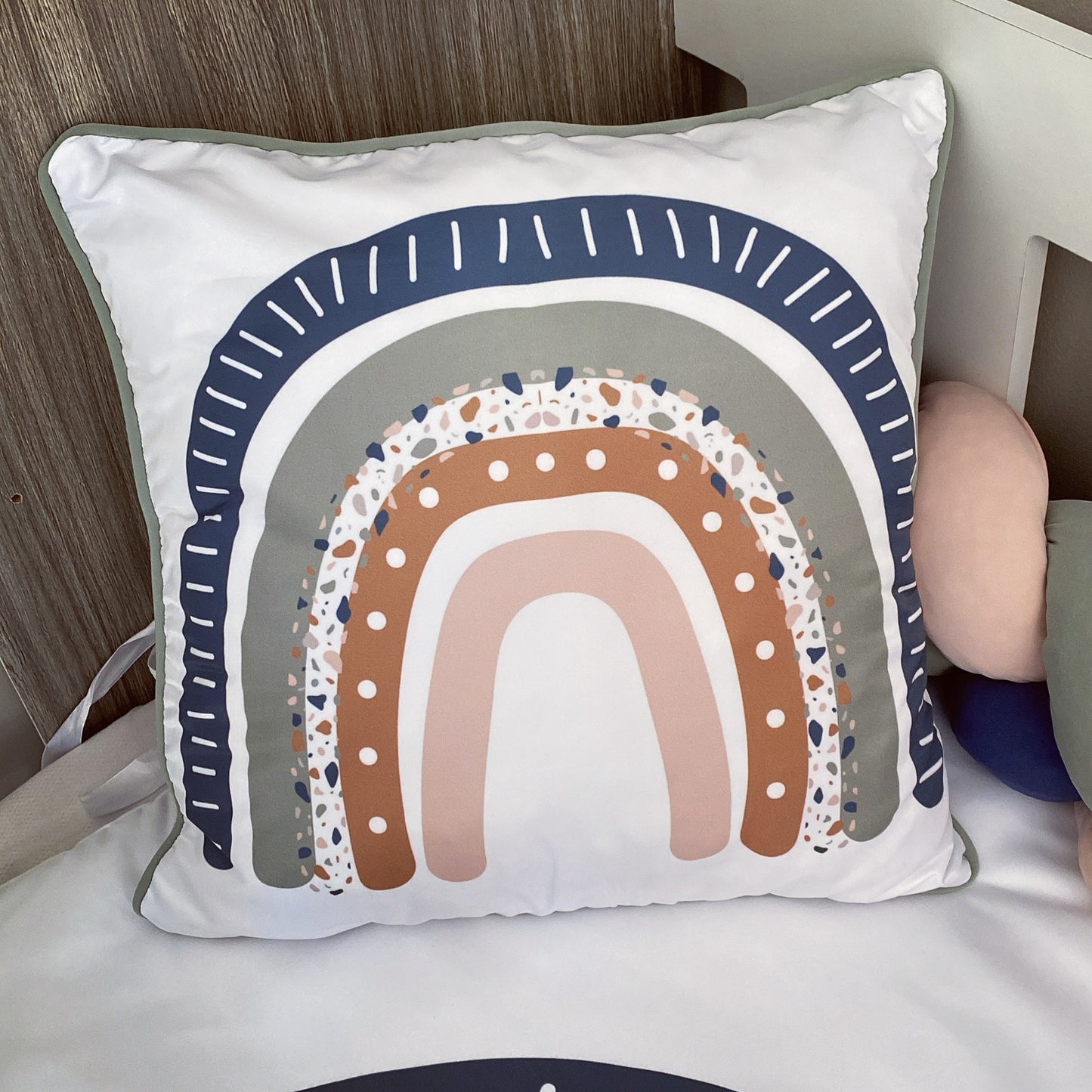 A square white throw pillow with a colorful rainbow design, resting on a white bed next to a wooden headboard.