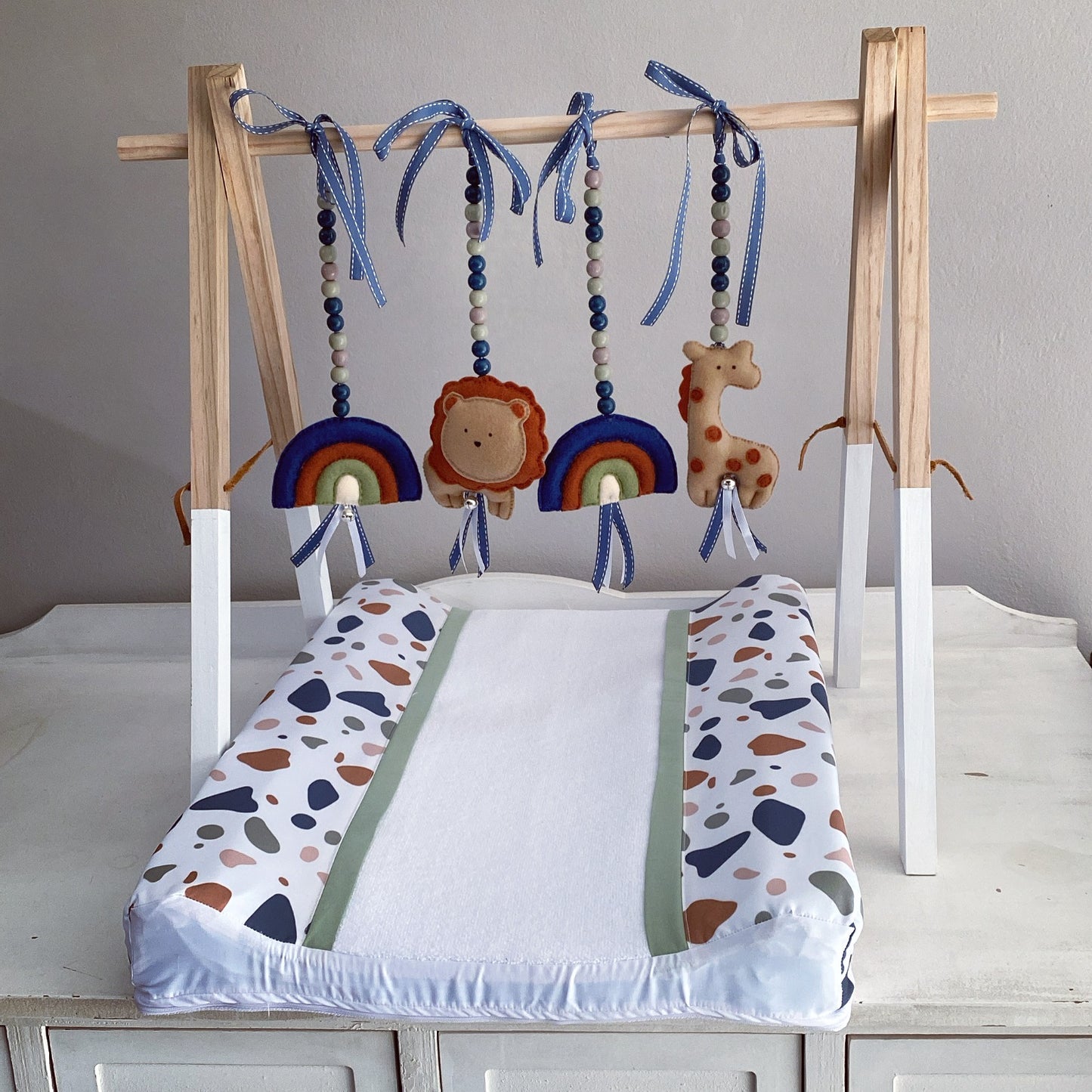 A baby changing pad with a wooden hanging toy bar, featuring colorful animal and rainbow toys, displayed on a white dresser against a neutral gray wall.