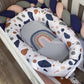 A baby bassinet cushion set in a white crib, featuring a padded oval bassinet cover with a white background and multicolor abstract shapes in navy, brown, and gray, surrounding a gray baby pillow with a rainbow design. | All Nursery Sets | Baby Bedding Sets South Africa | Soft Spot Kids | South Africa