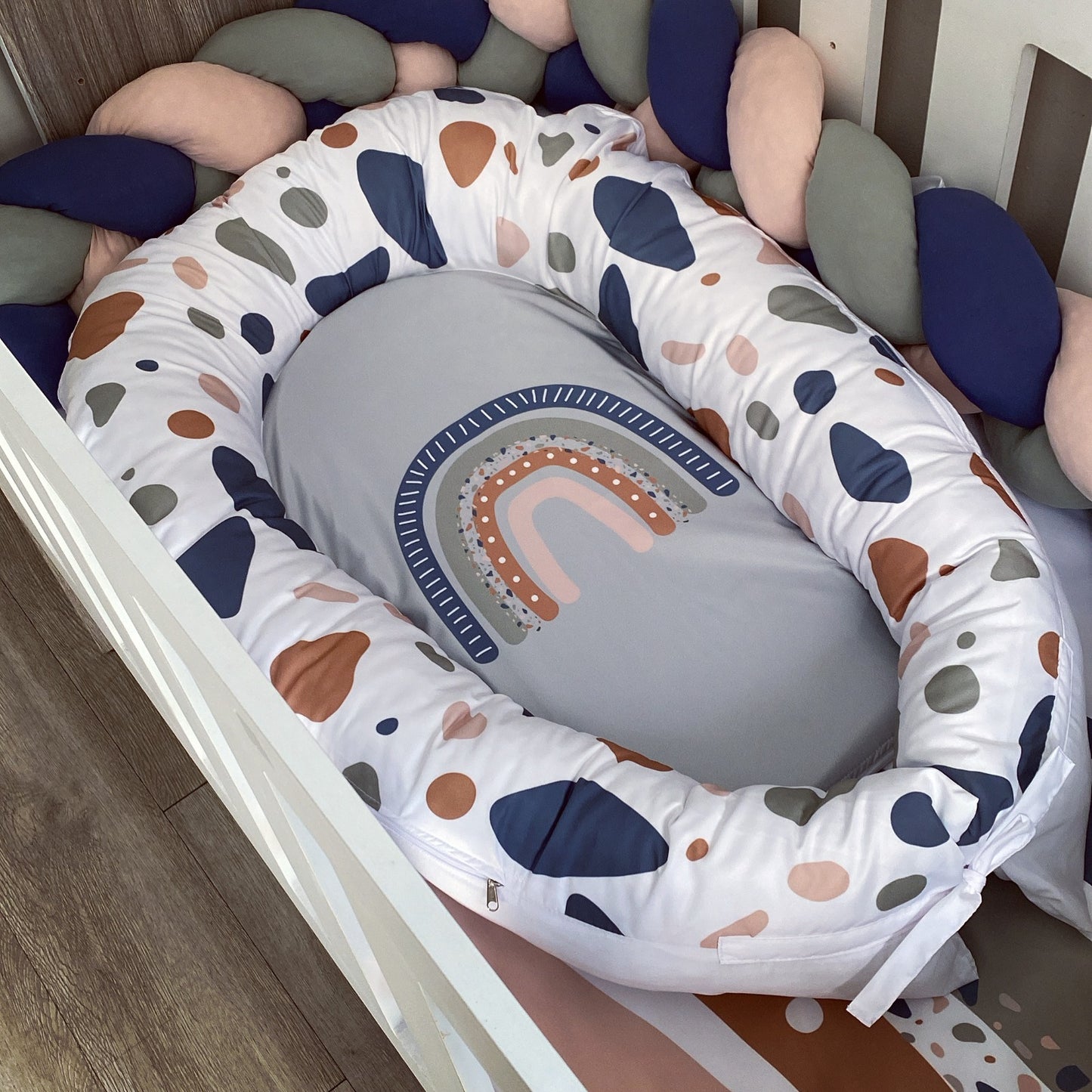 A baby bassinet cushion set in a white crib, featuring a padded oval bassinet cover with a white background and multicolor abstract shapes in navy, brown, and gray, surrounding a gray baby pillow with a rainbow design.