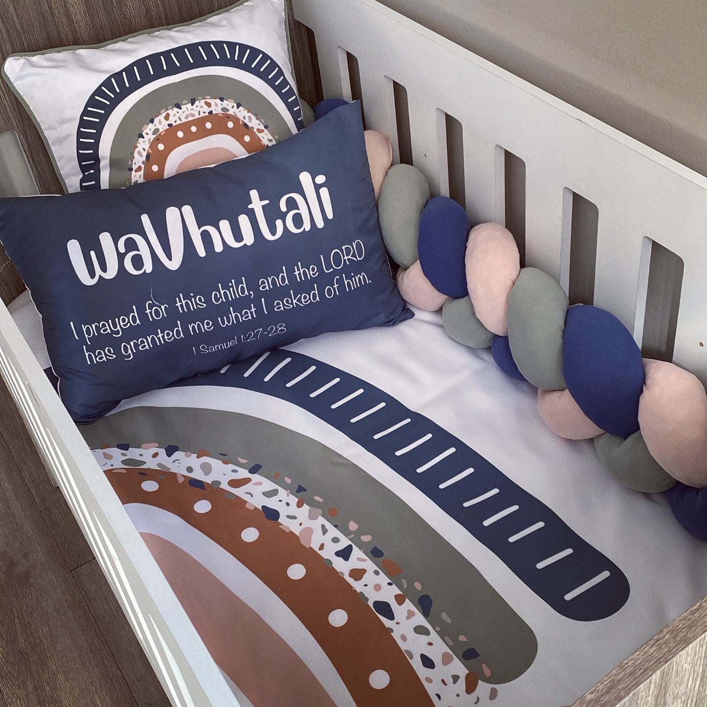 A styled nursery crib setup featuring a personalized navy blue pillow with white text reading 'waVutali I prayed for this child, and the LORD has granted me what I asked of him', a matching printed bumper cushion, and a chunky braided crib bumper in navy, blush pink, and gray.