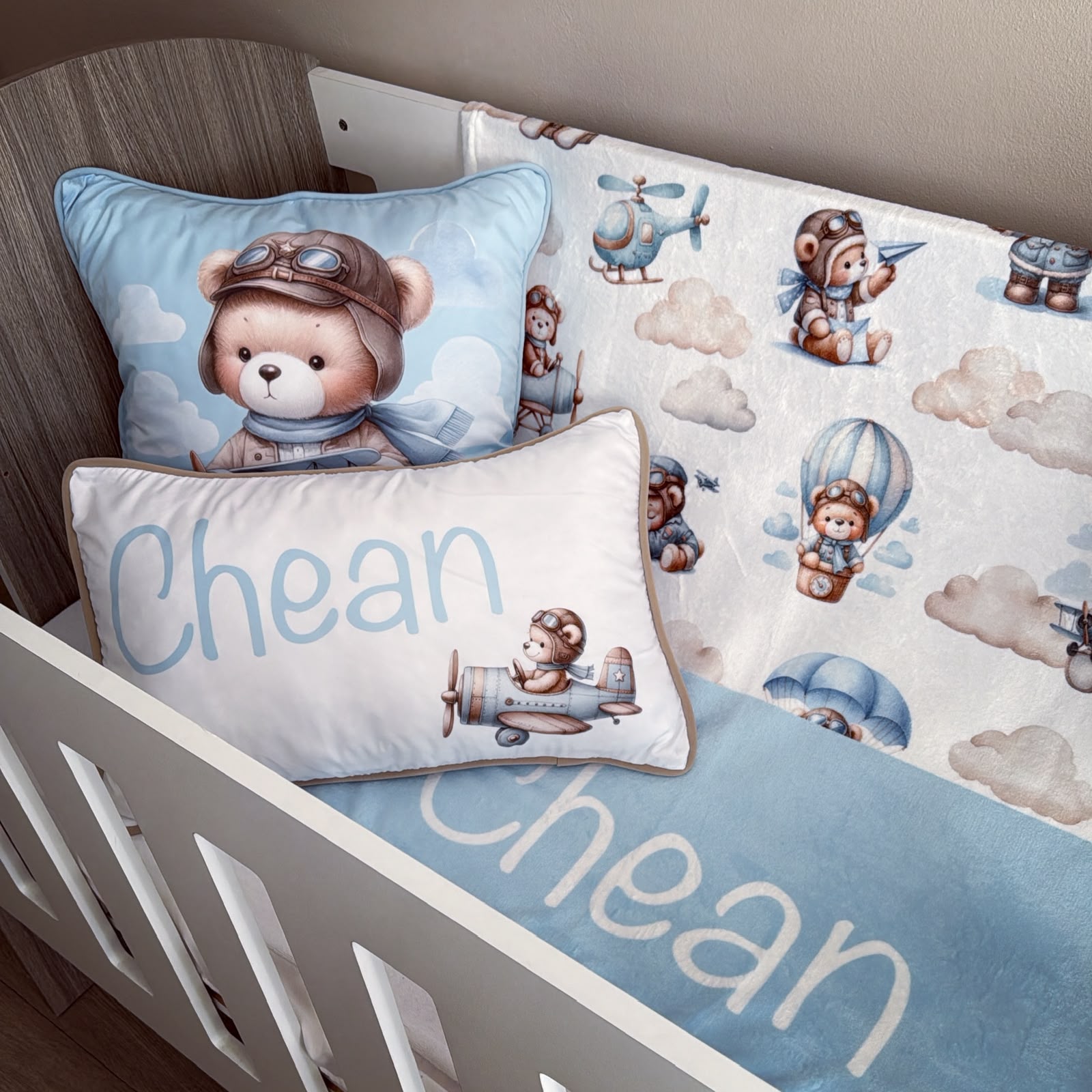 A baby crib bedding set featuring a white and light blue quilt with cartoon teddy bear aviator prints, a matching blue fitted sheet, and two personalized pillows with the name 'Chean' in white script.