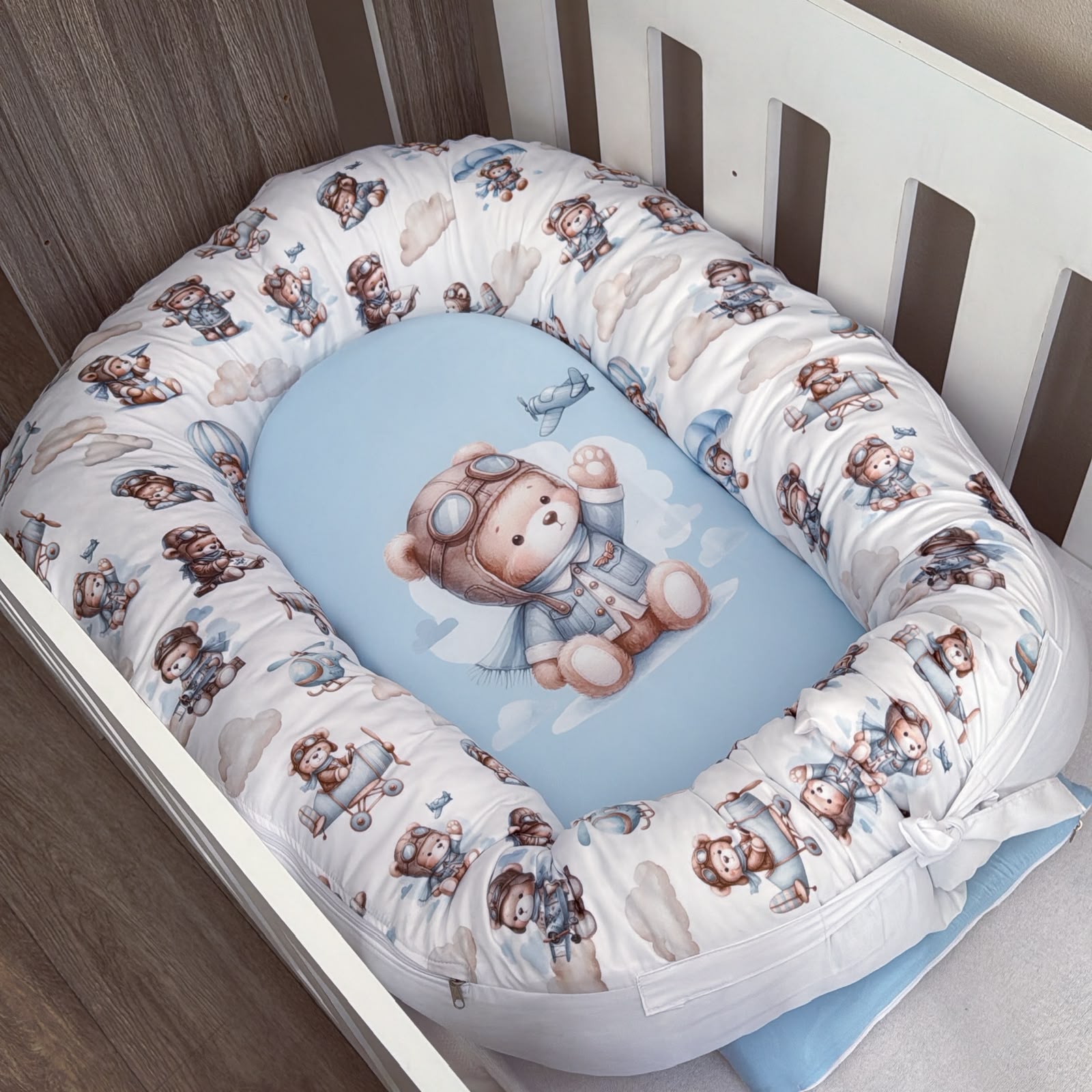 A baby crib bedding set featuring a white bassinet bumper pad with a brown teddy bear and airplane pattern, surrounding a light blue oval mattress topper with a matching teddy bear illustration.