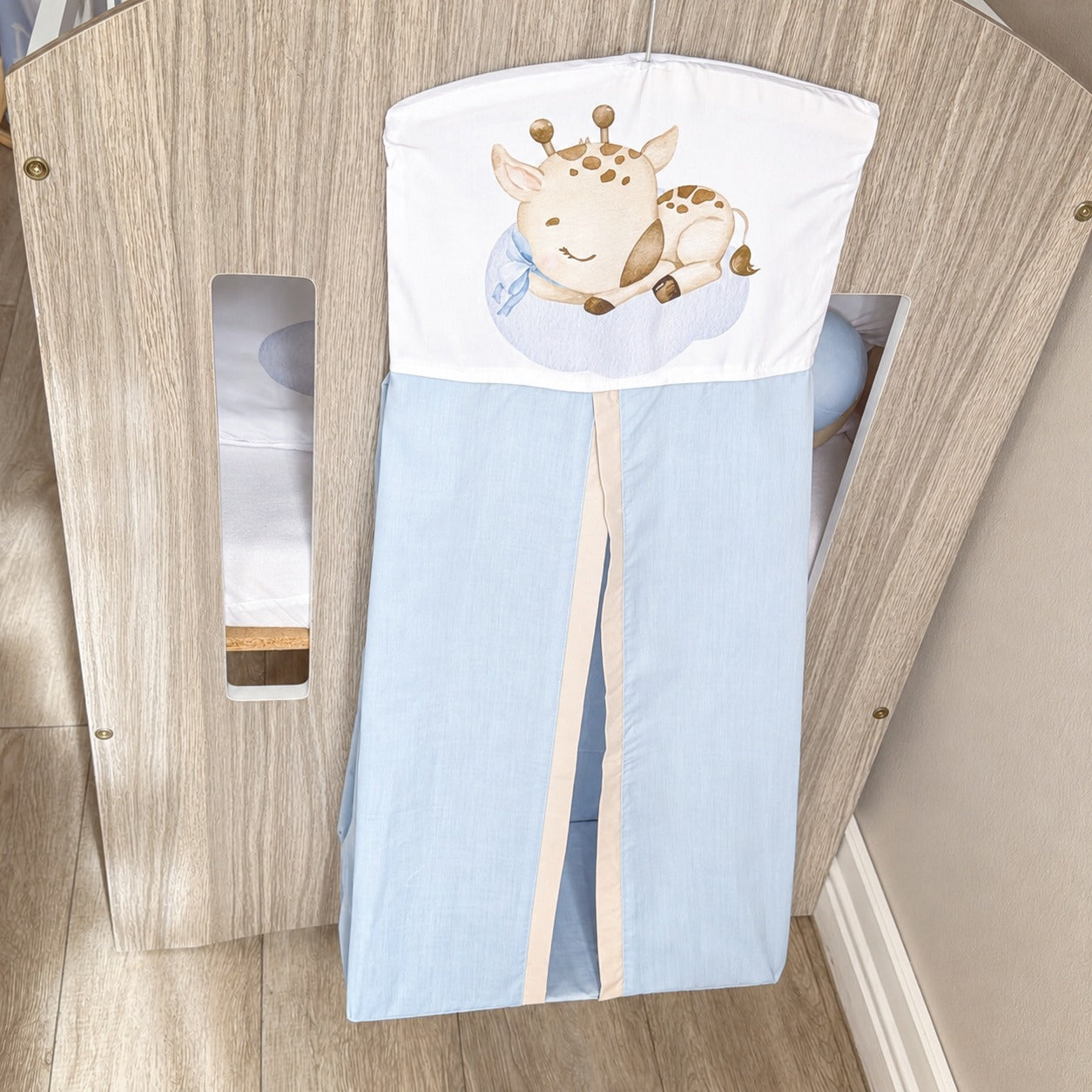 A baby crib nappy holder with a light blue body and a white top panel featuring a cartoon sleeping giraffe, shown draped over a wooden crib.