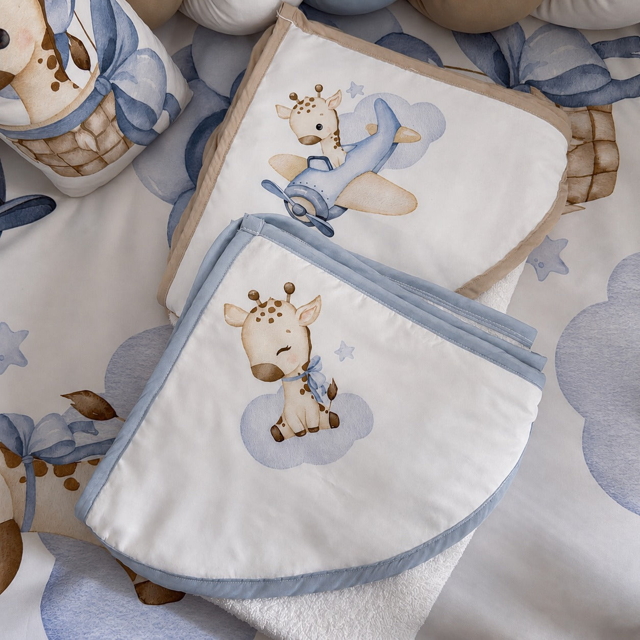 A styled flat-lay of baby bedding and hooded towels a whimsical giraffe and airplane print in soft blue, beige, and white tones. The set includes a folded blanket, a matching pillow or cushion, and a cylindrical container, all arranged on a coordinating patterned sheet.