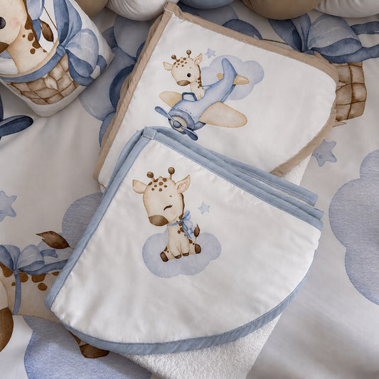 A styled flat-lay of baby bedding and hooded towels a whimsical giraffe and airplane print in soft blue, beige, and white tones. The set includes a folded blanket, a matching pillow or cushion, and a cylindrical container, all arranged on a coordinating patterned sheet.