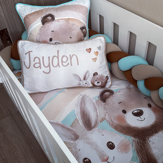 A personalized baby bedding set in a white crib, featuring a printed comforter and pillowcase with cartoon bear and bunny illustrations, a custom name pillow reading 'Jayden', and a chunky braided bumper in brown, beige, and light blue.