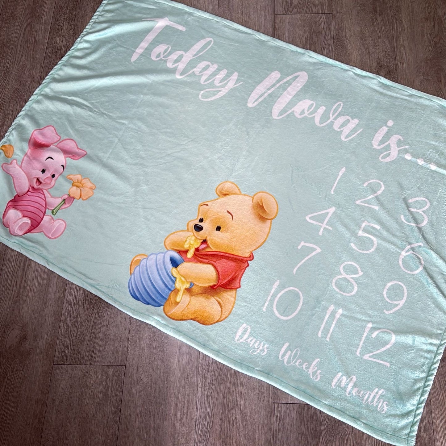Baby milestone blanket with cartoon characters and numerical text on a wooden floor.