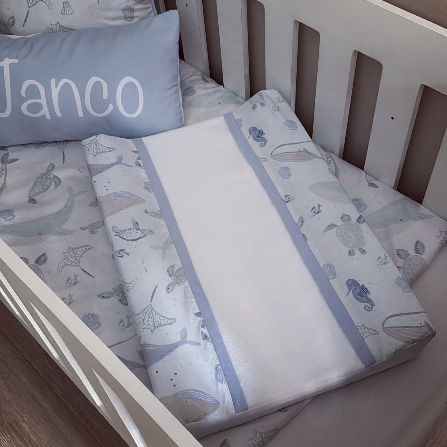 A baby bedding set in a white crib, featuring a duvet cover and fitted sheet with a light blue and gray sea life pattern, a matching pillow, and a solid light blue accent strip.