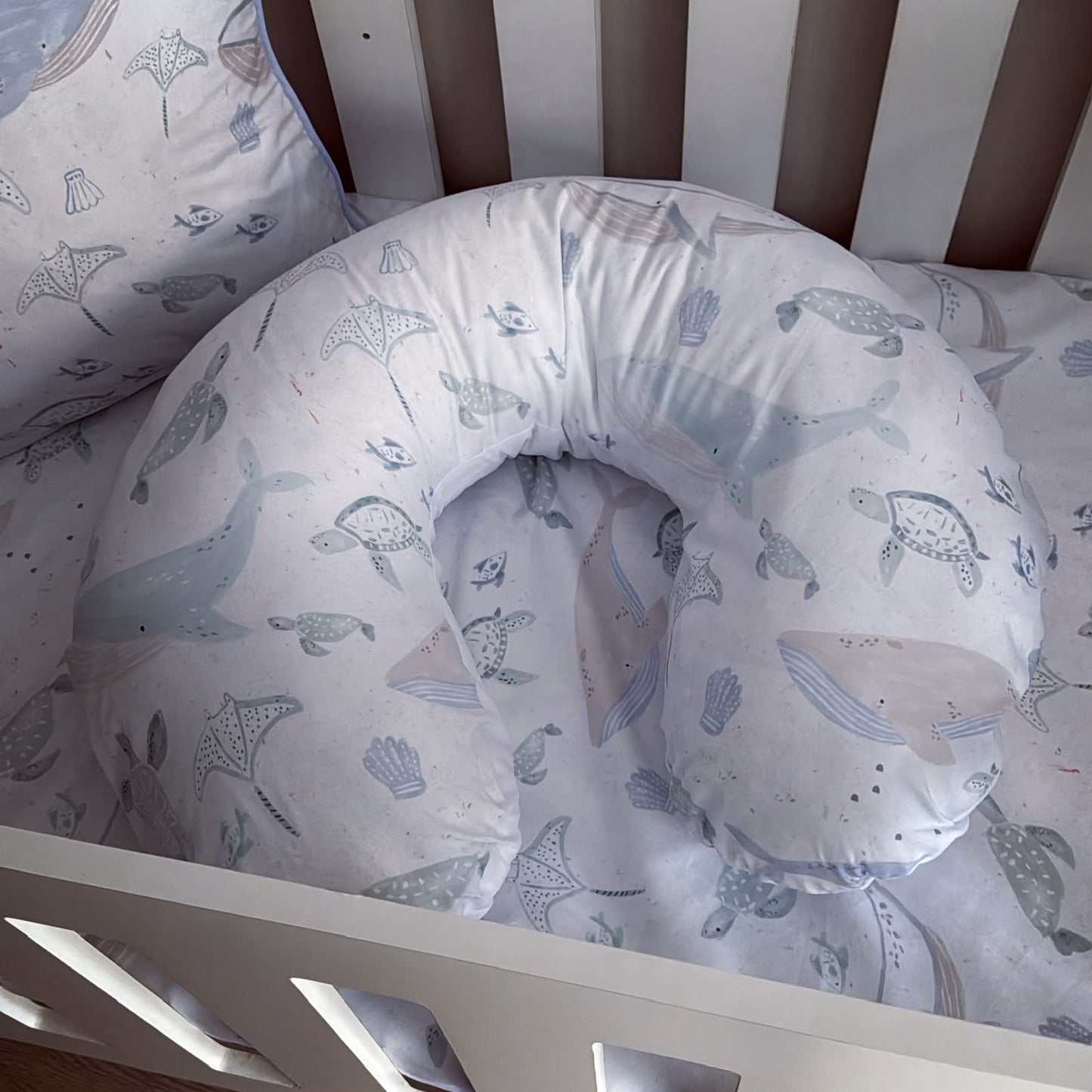 A U-shaped baby support pillow with matching crib bedding featuring a light blue and beige whale, turtle, and seashell pattern, shown inside a white wooden crib.