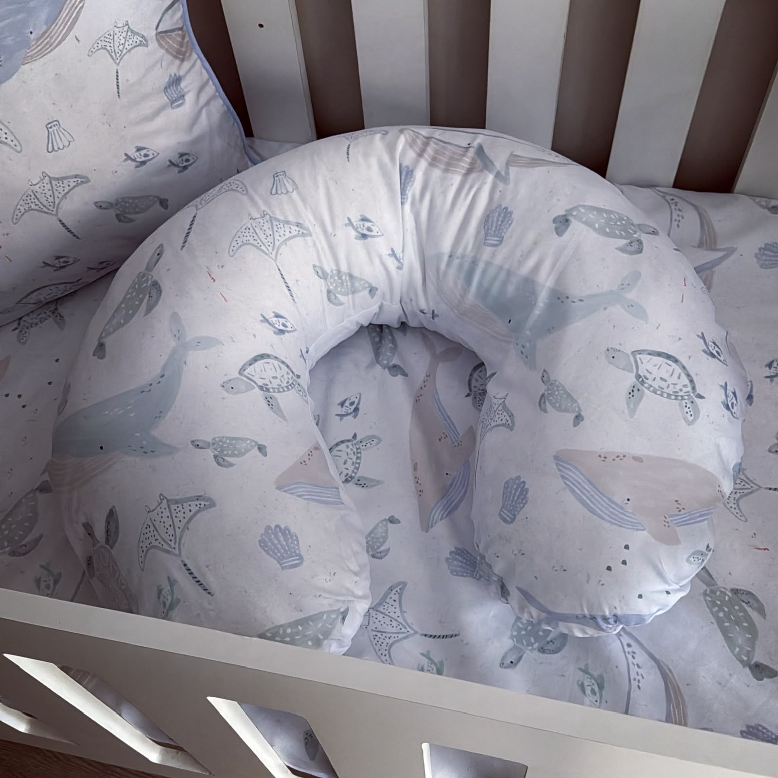 A U-shaped baby support pillow with matching crib bedding featuring a light blue and beige whale, turtle, and seashell pattern, shown inside a white wooden crib.