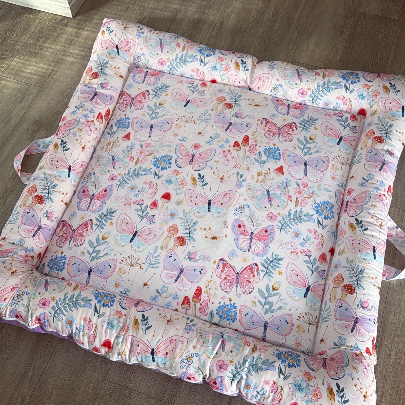 A square padded baby floor doughnut mat with a ruffled border, featuring a colourful butterfly and floral print on a white background, laid out on a wooden floor.