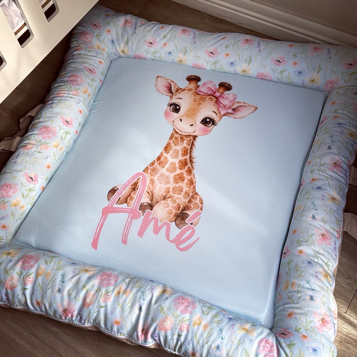 A square baby pet bed with a light blue center featuring a watercolor giraffe and the name 'Ame' in pink script, surrounded by a floral-patterned border.