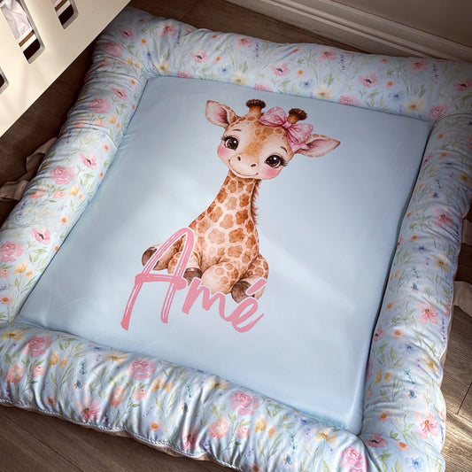 A square baby pet bed with a light blue center featuring a watercolor giraffe and the name 'Ame' in pink script, surrounded by a floral-patterned border.