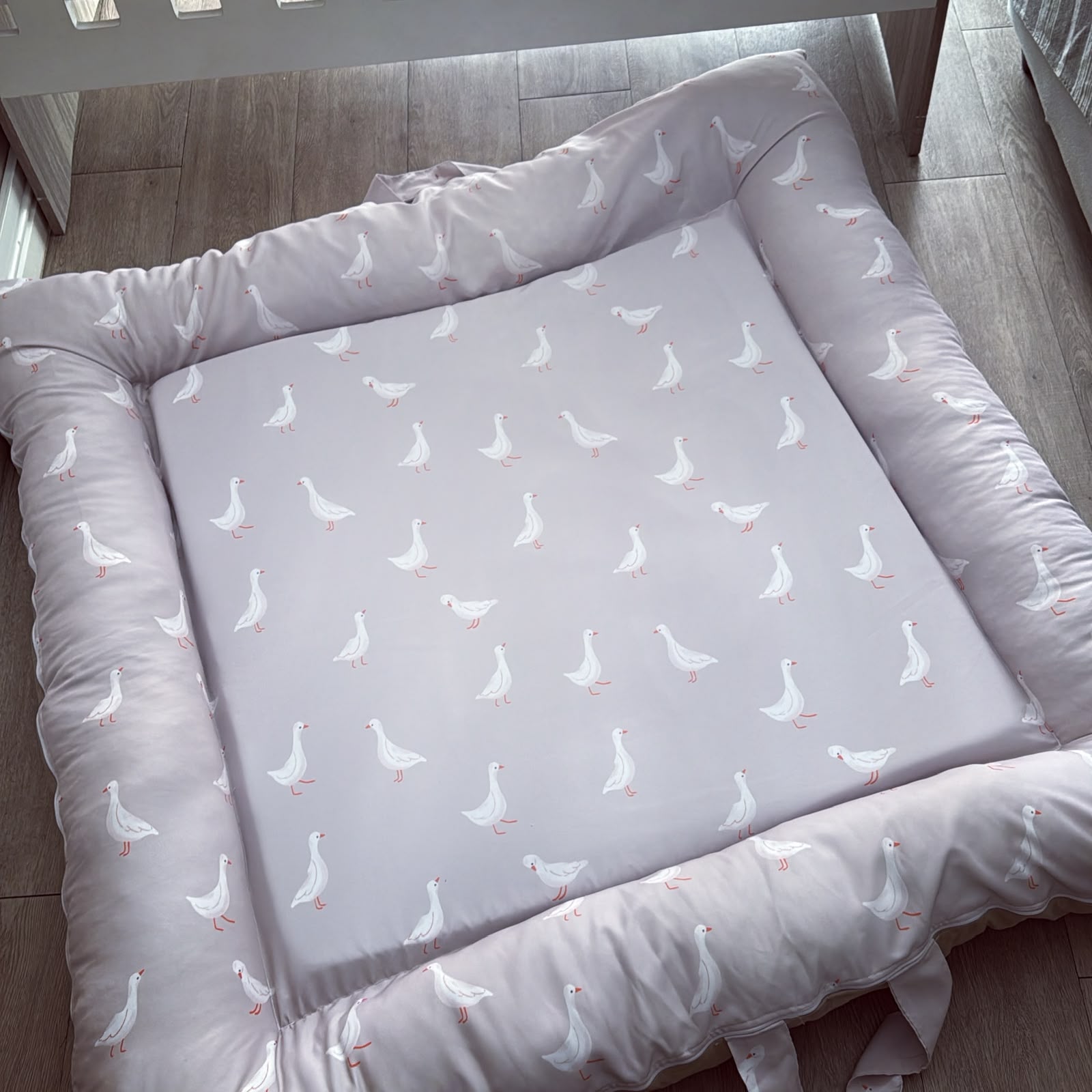 A padded baby mattress pad with a matching padded border, featuring a light gray center printed with a repeating pattern of white geese with orange beaks and feet, shown on a wooden floor.
