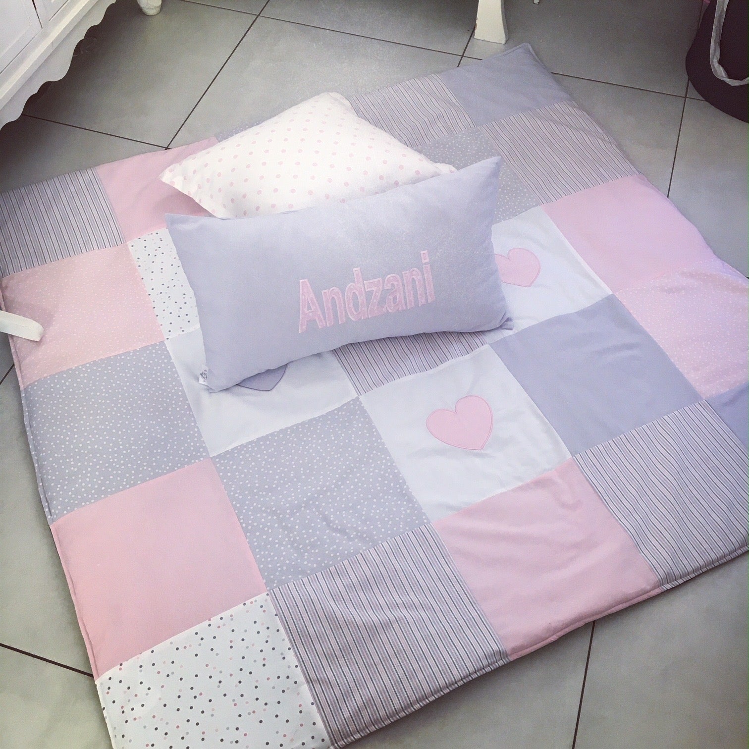 Patchwork blanket with pink and gray squares and a heart design, featuring a pillow with 'Andzani' on a tiled floor.