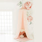 A pink hanging tent canopy with floral designs, set up in a room with a white background. It is positioned above a cream-colored feeding chair with a rose gold accent. | Baby Canopy Nets | Cot Mosquito Nets | Soft Spot Kids | South Africa