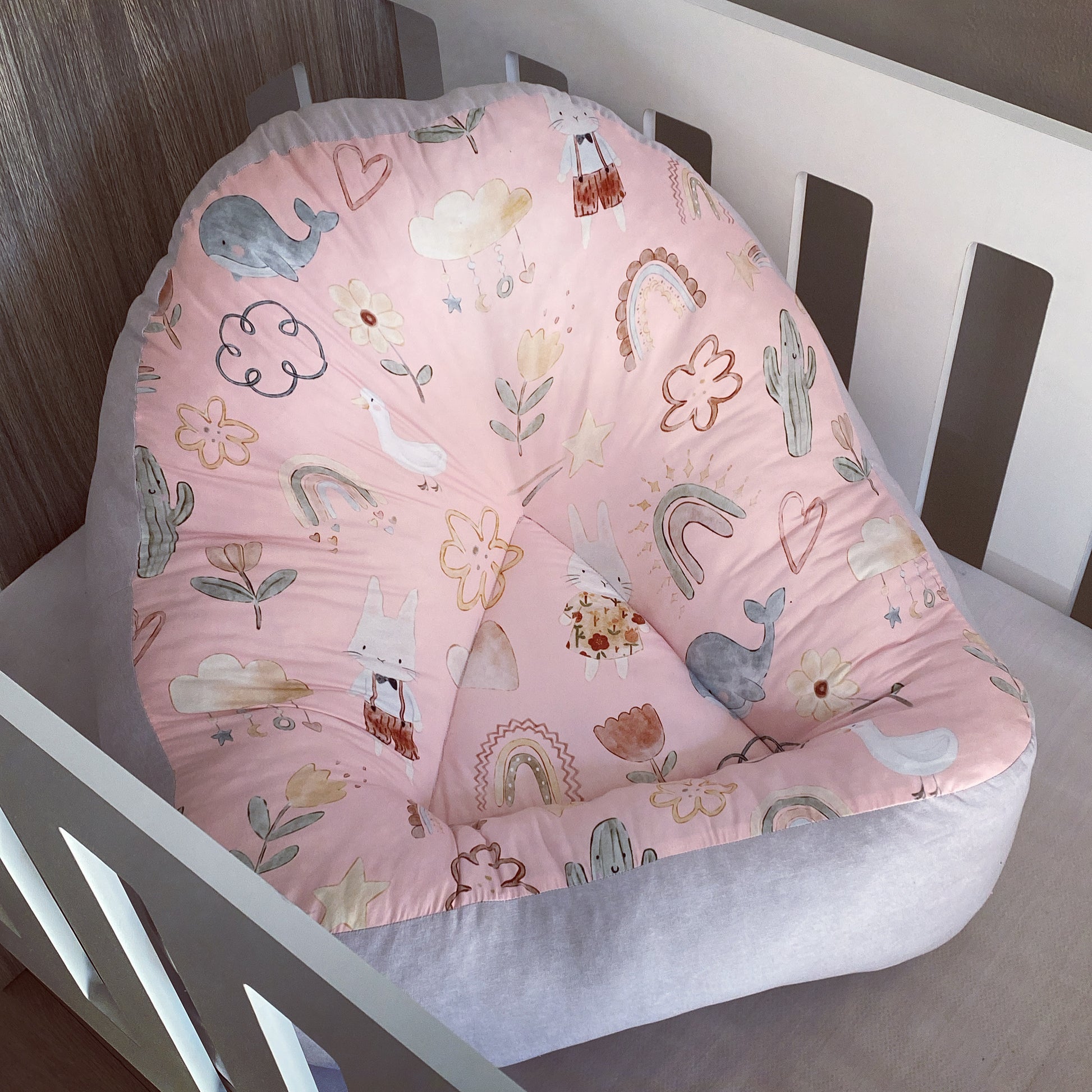 A pink boho-style baby lounger with a pattern of animals and plants, placed in a wooden crib.